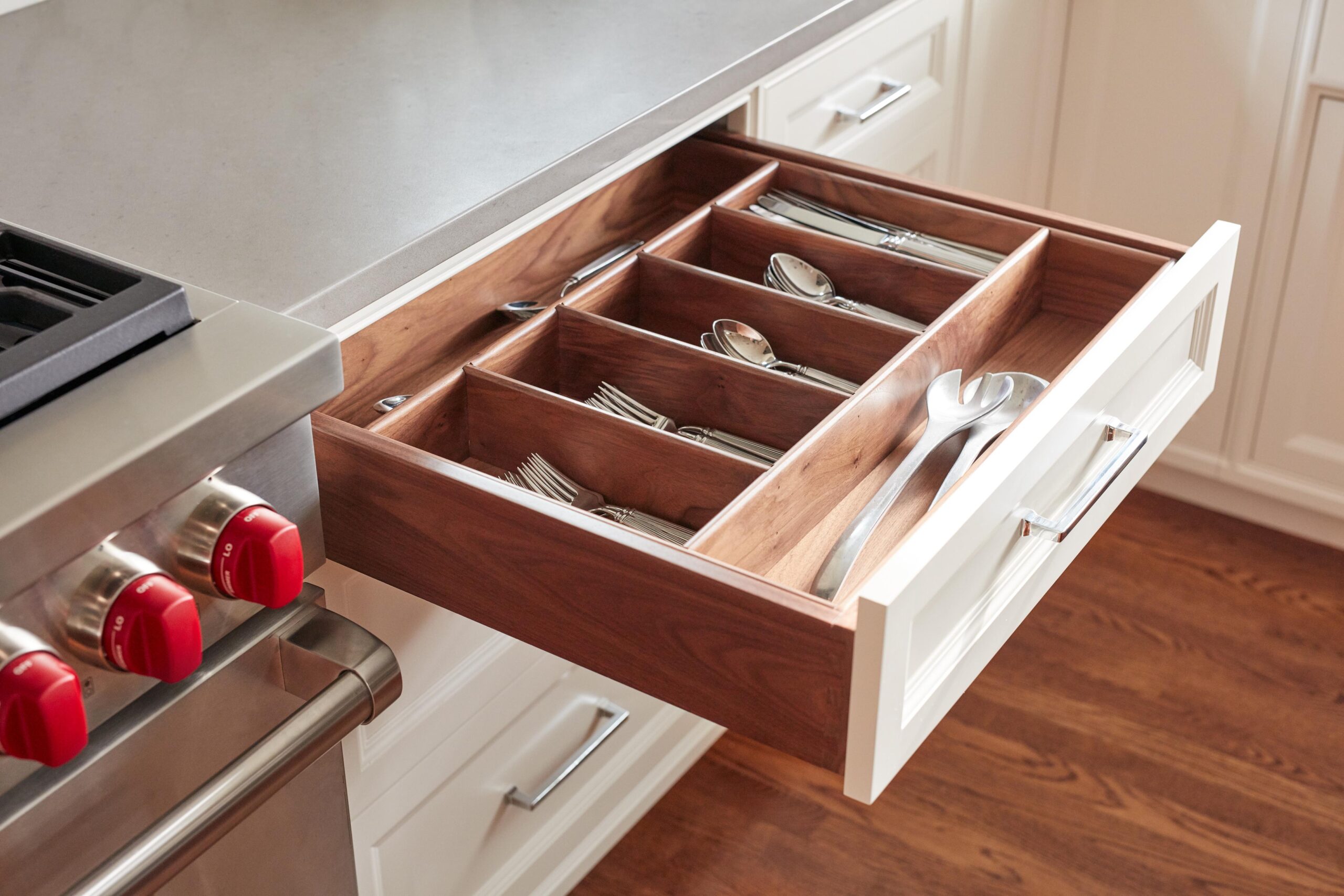 A wooden kitchen drawer with dividers is open, displaying organized silverware including forks, knives, spoons, and serving utensils next to a stainless steel stove.
