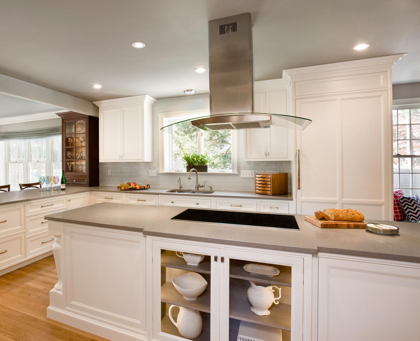 Modern kitchen with white cabinets, a gray countertop island, built-in stovetop, stainless steel range hood, and natural light from multiple windows.