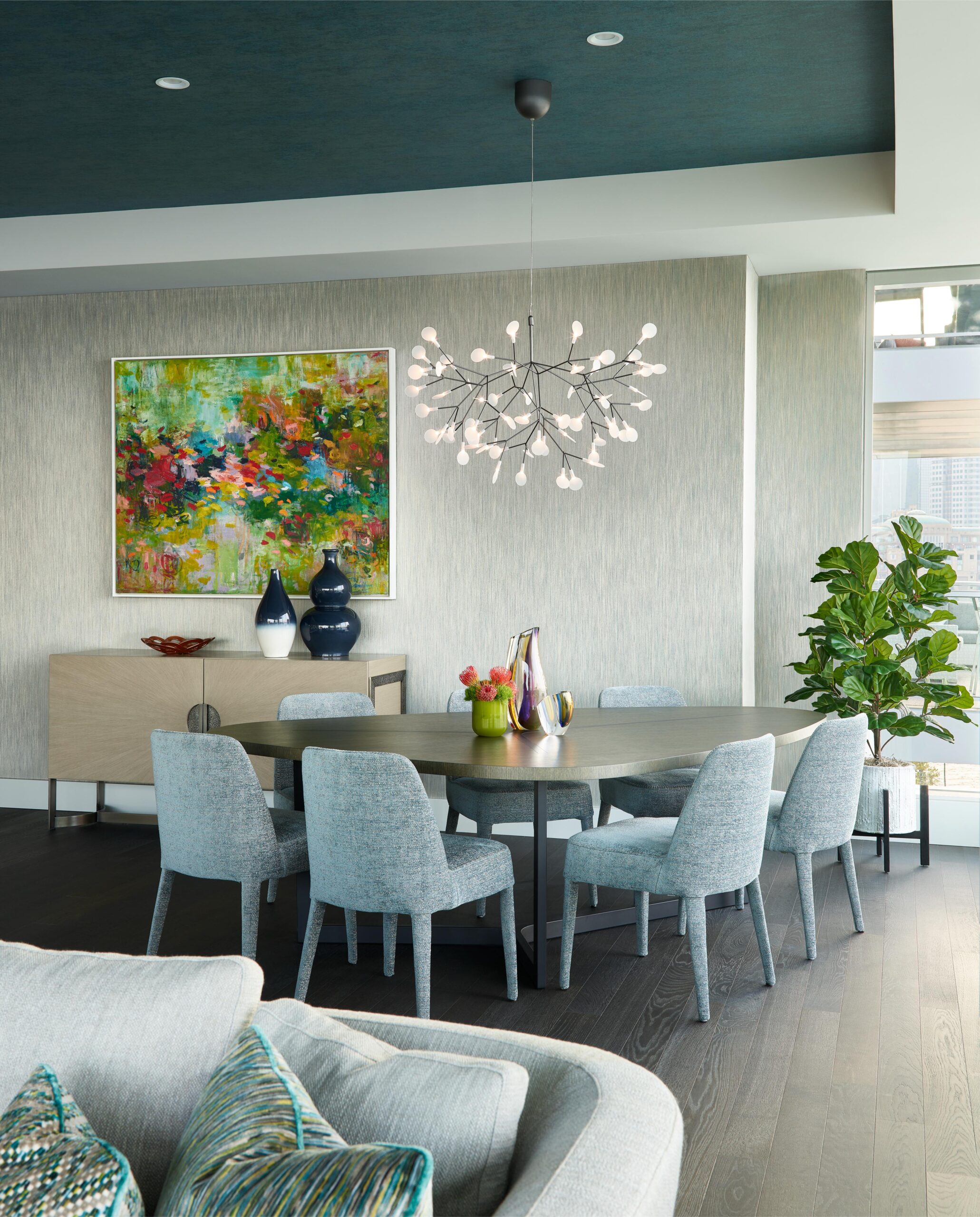 Modern dining area with a round table, eight gray chairs, a sideboard, abstract wall art, decorative vases, a potted plant, and a contemporary chandelier.