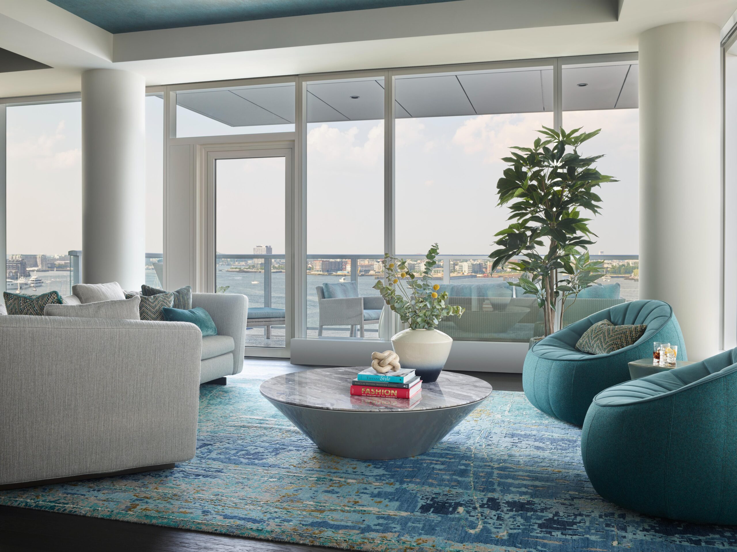 Modern living room with teal chairs, a grey sofa, round marble coffee table with books and vases, large windows, a potted plant, and a view of the city and water outside.