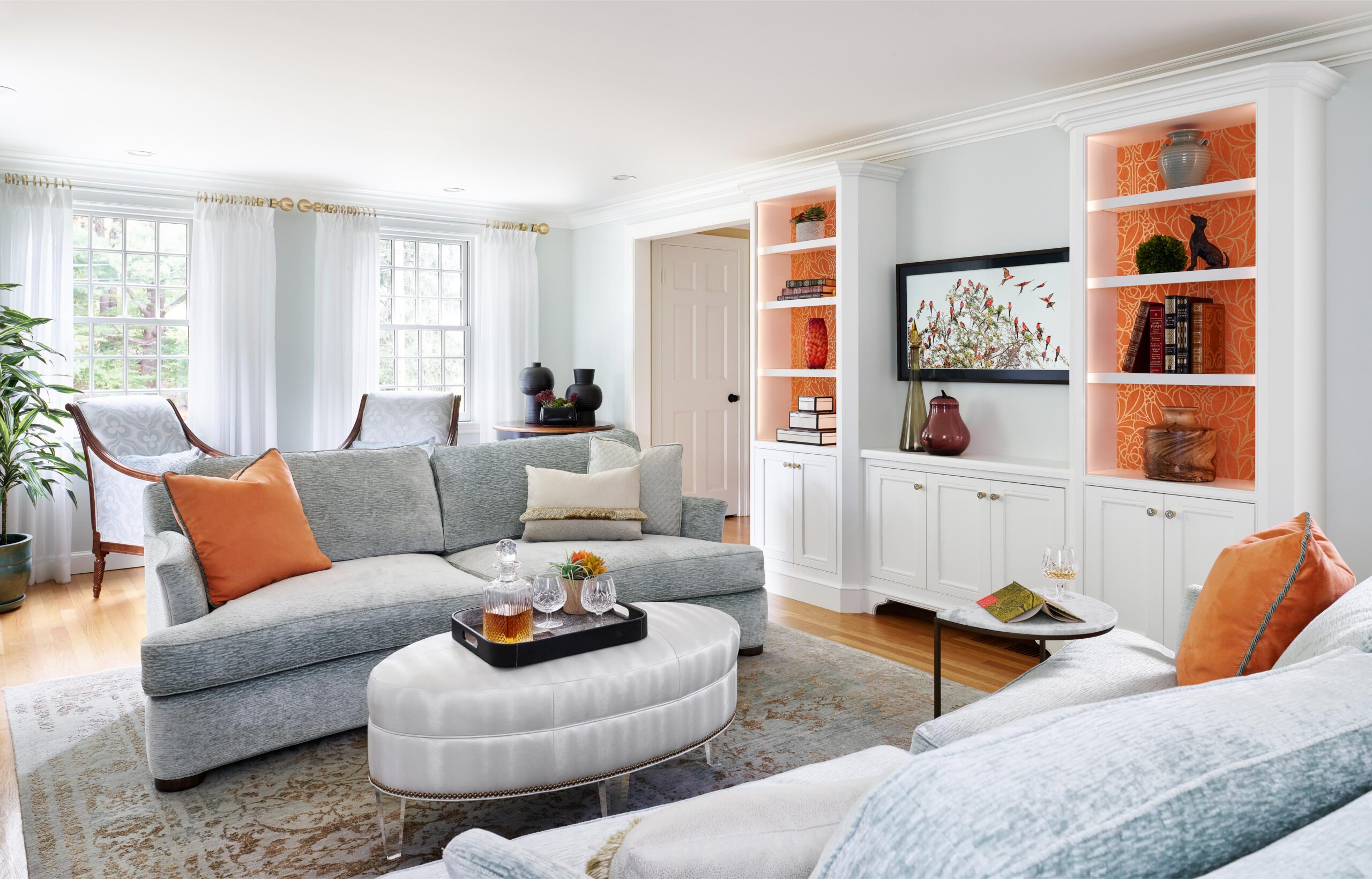 A bright living room with light blue sofas, orange accent pillows, a white ottoman, built-in shelves with orange backing, books, and decorative items, and large windows with white curtains.