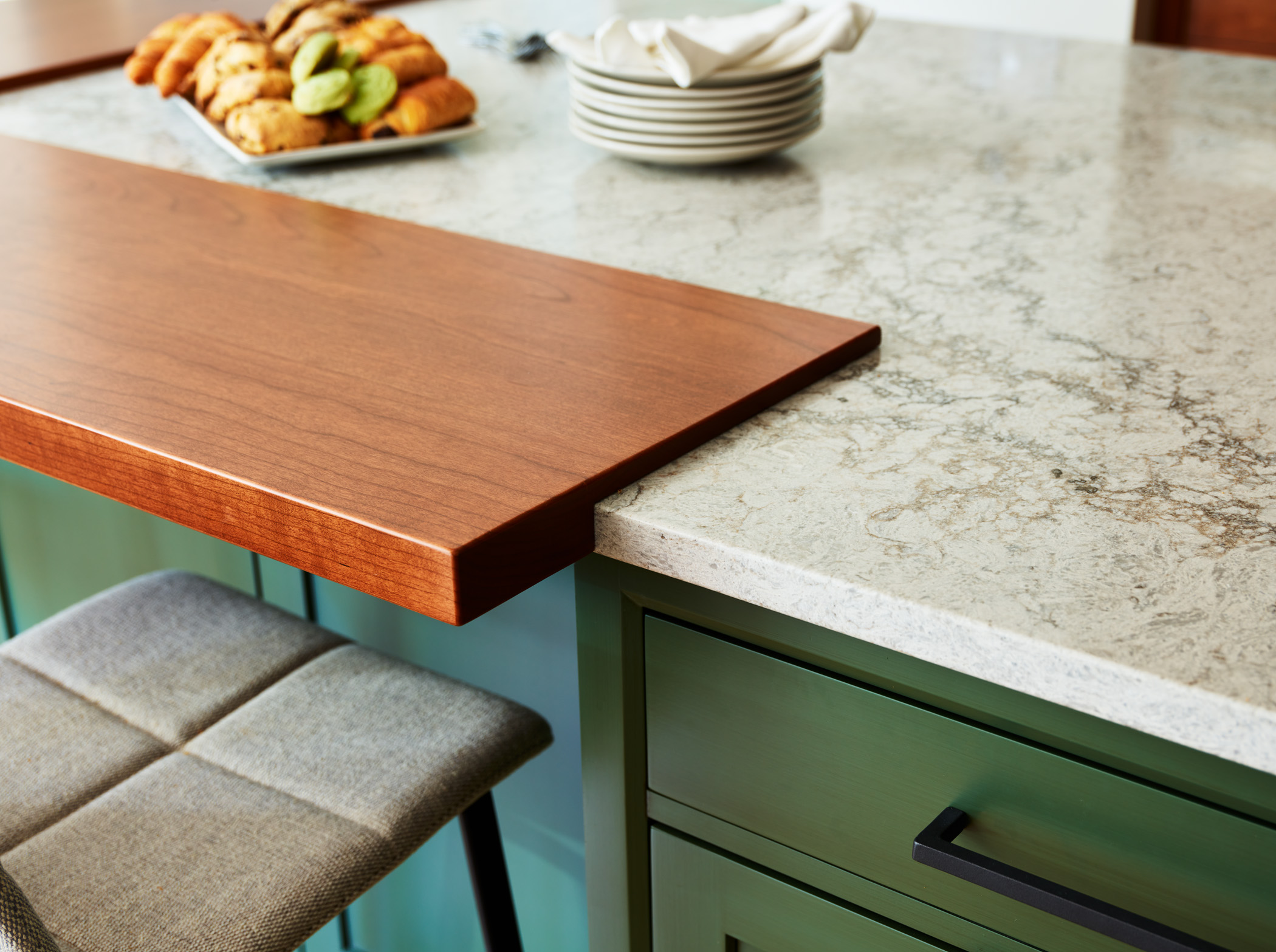 A close-up of a kitchen island with a wooden breakfast bar, marble countertop, green cabinetry, and elegant touches—a plate of pastries, stacked dishes, and a cushioned stool—reflecting luxury interior design by an expert Interior Designer in Massachusetts.