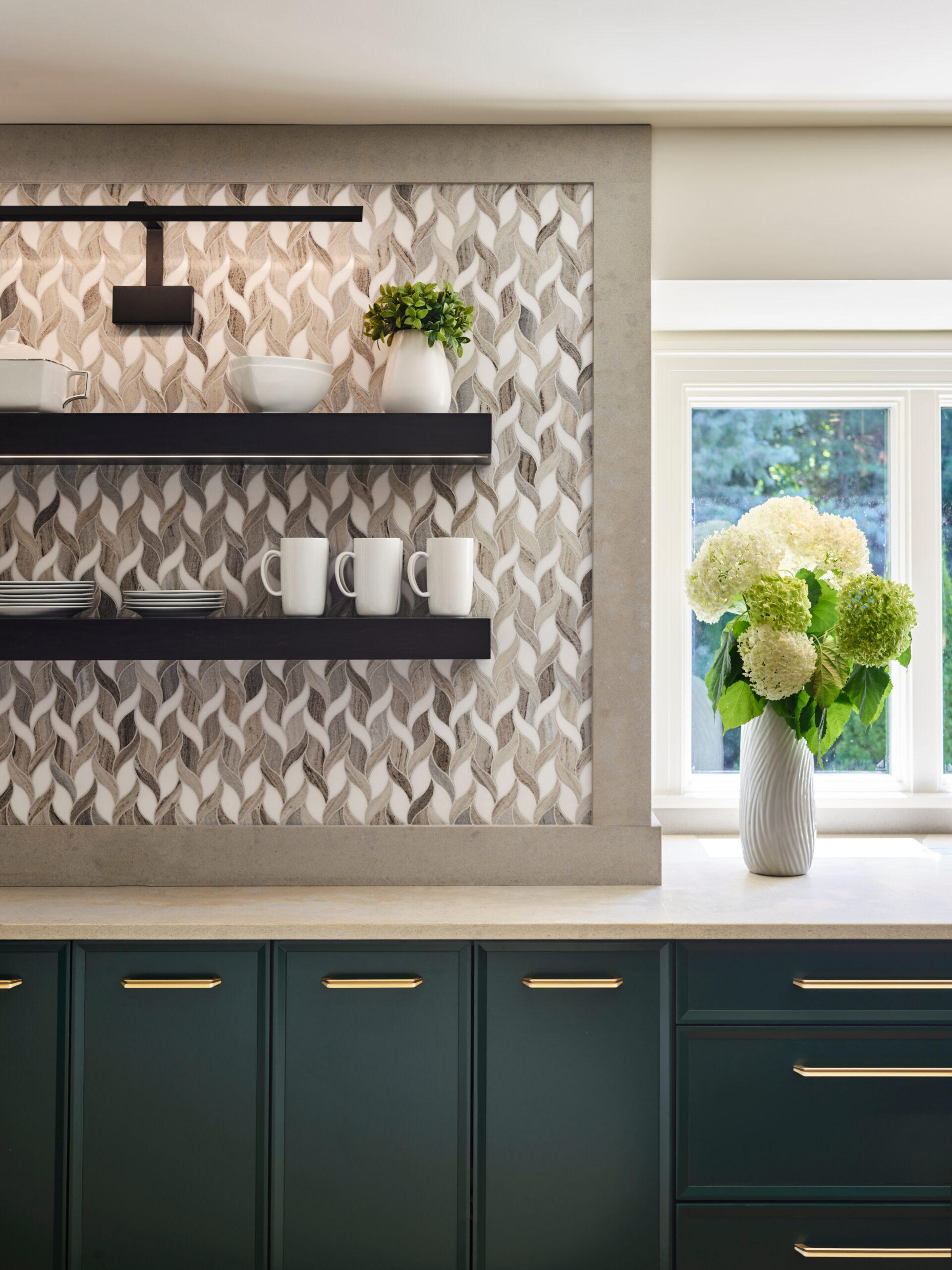 Modern kitchen with green cabinets, gold handles, and open shelves holding dishes and mugs. A geometric tile backsplash adds flair, while a vase of hydrangeas brightens the counter—a stunning example of luxury interior design near a sunlit window.