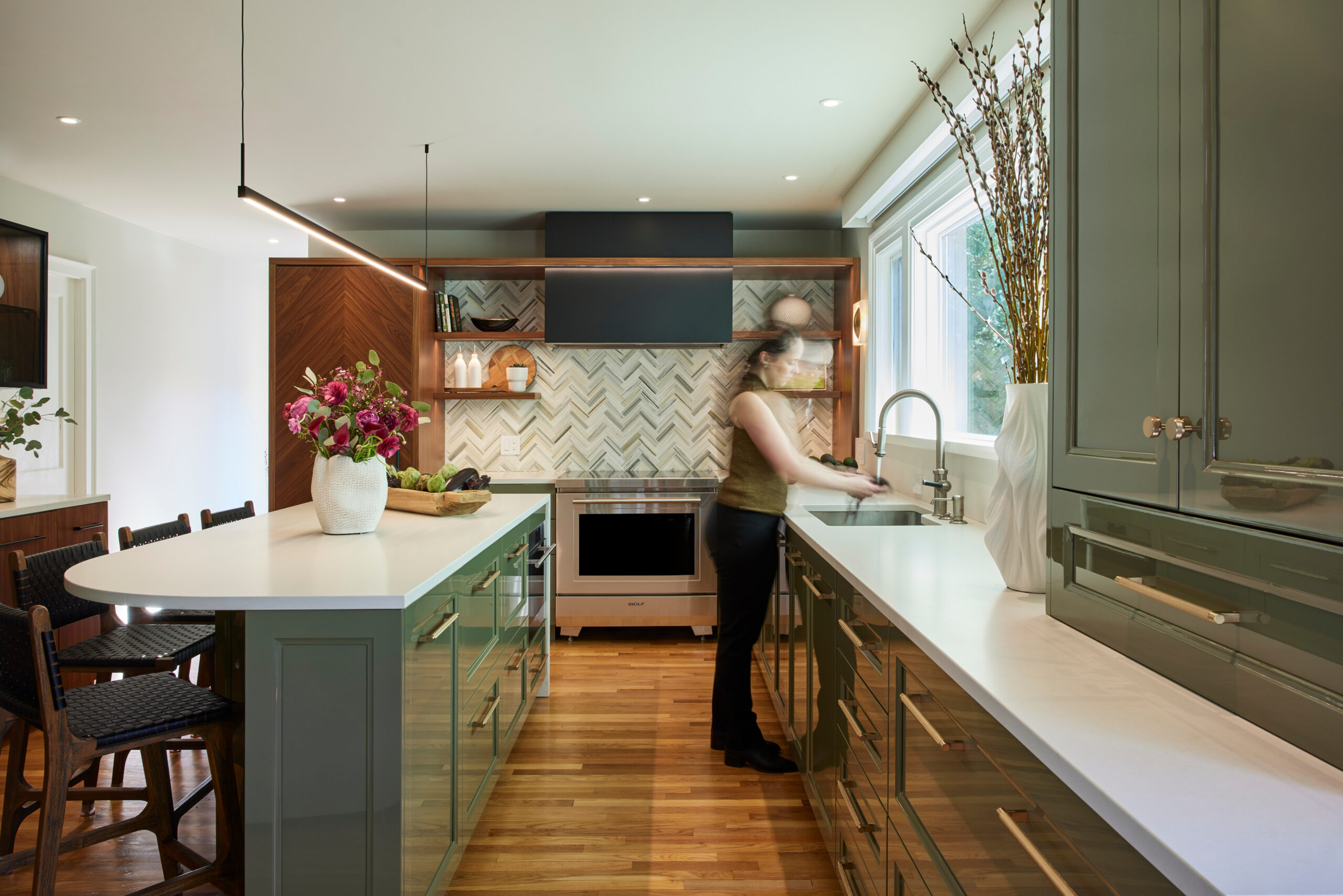 A person stands at a sink in a modern kitchen with green cabinets, white countertops, a center island, and herringbone tile backsplash—an inspiring example of luxury interior design by an interior designer in Massachusetts.
