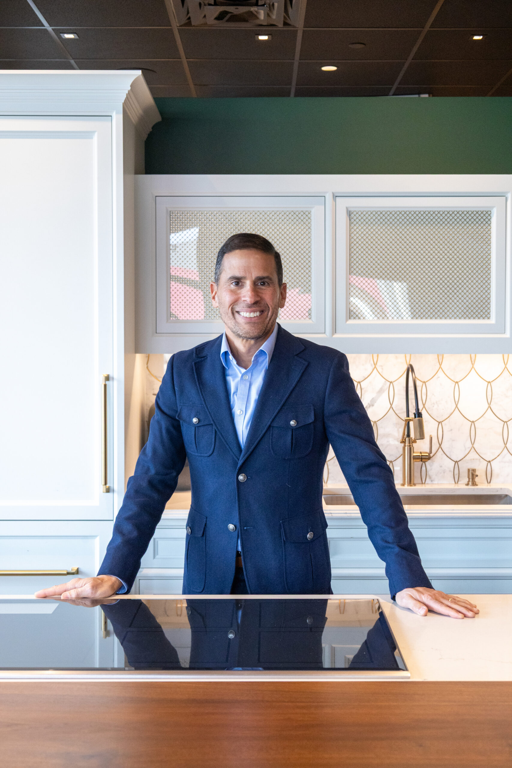 A man in a blue blazer stands smiling in a modern kitchen, featuring luxury interior design with white cabinets, a tiled backsplash, and a cooktop in front of him.