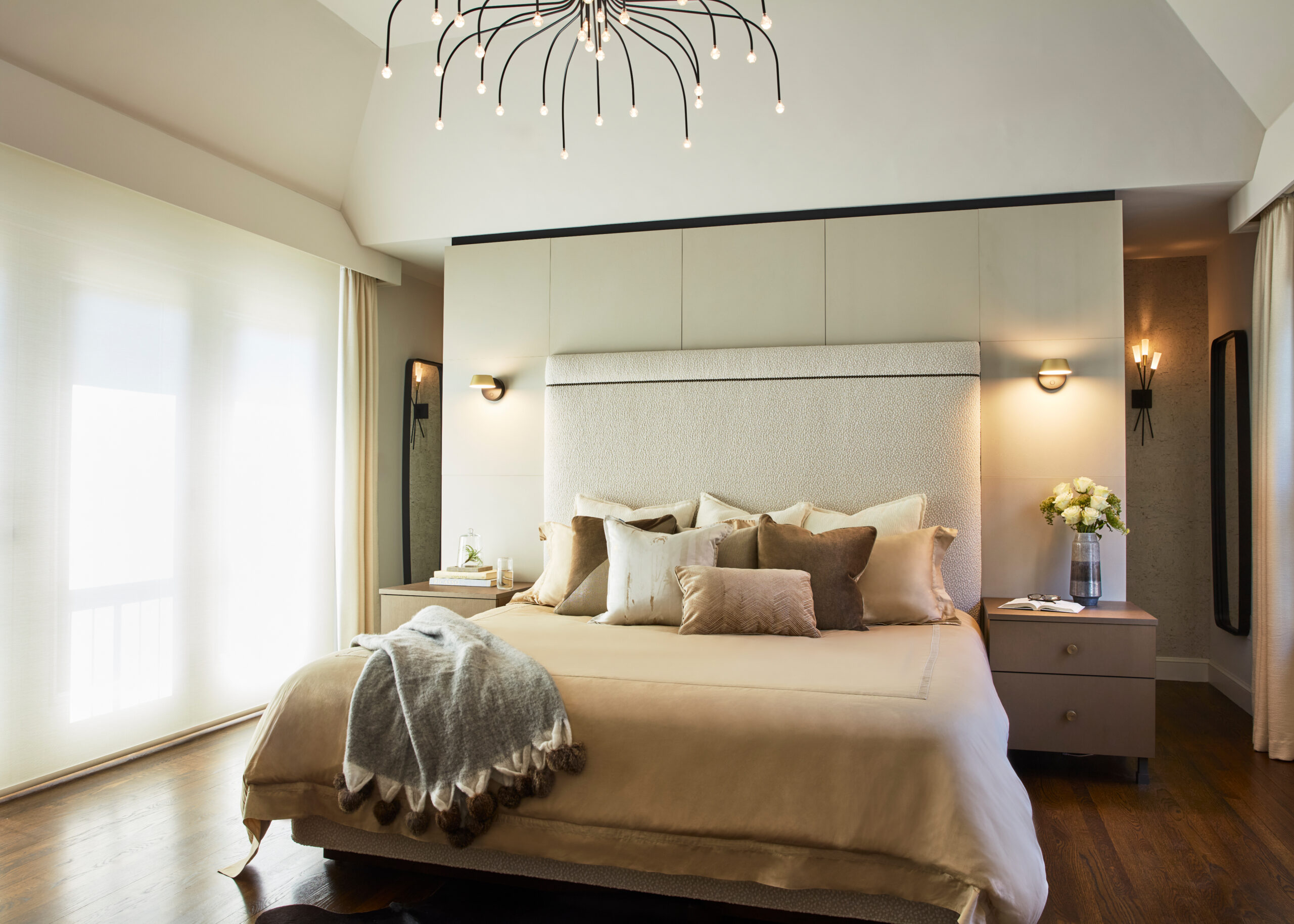 A modern bedroom with a large upholstered bed, multiple pillows, bedside tables with lamps, and a contemporary chandelier showcases luxury interior design. Light streams in through sheer curtains, creating an inviting atmosphere.