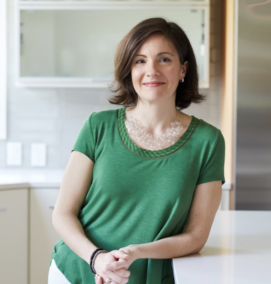 Woman with short brown hair, an Interior Designer from Massachusetts, wears a green shirt and clear beaded necklace as she stands and smiles in a modern, bright kitchen featuring luxury interior design.