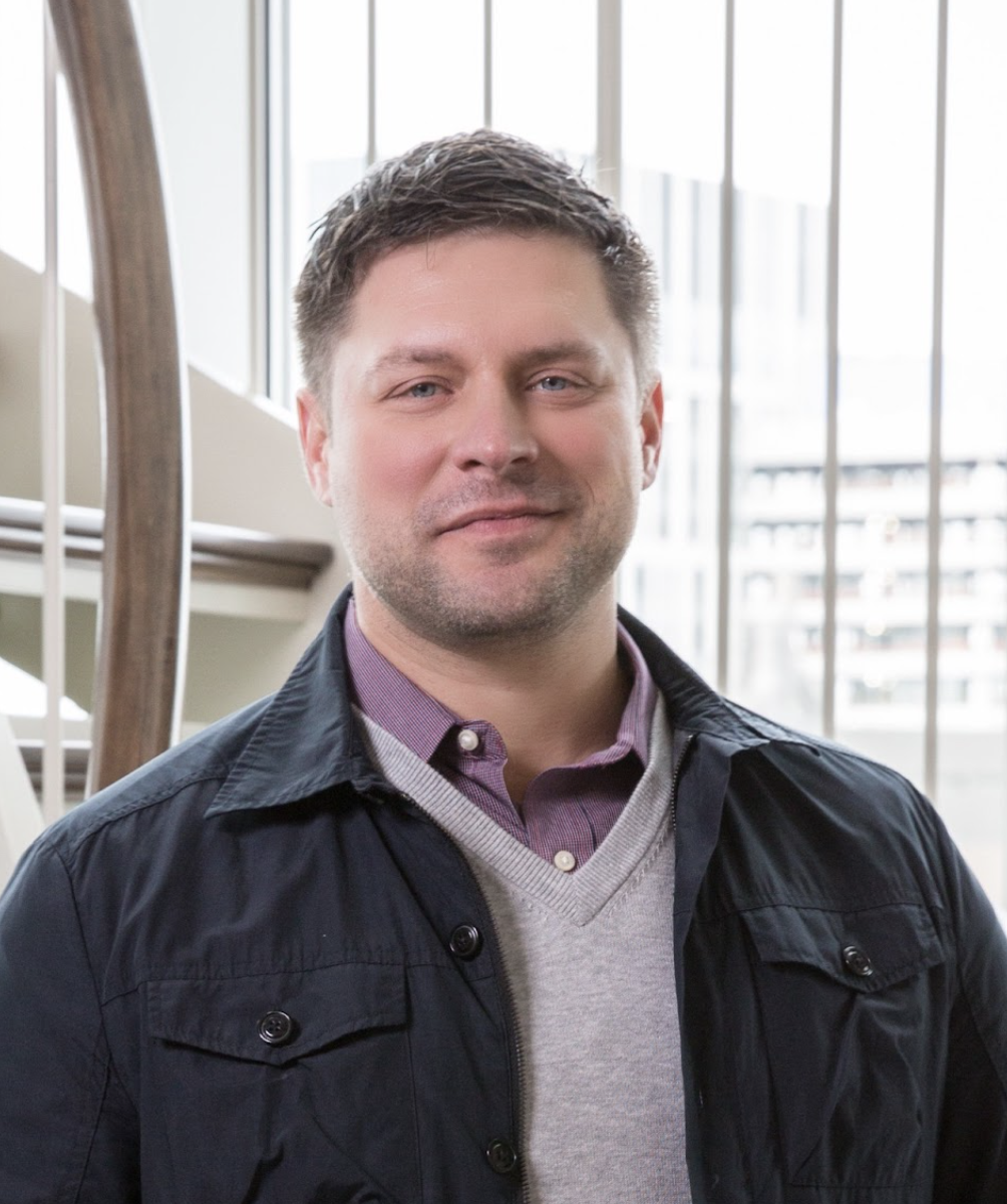 A man with short brown hair and stubble, wearing a dark jacket over a gray sweater and collared shirt, stands indoors in front of large windows with vertical blinds in a space reflecting Luxury Interior Design.