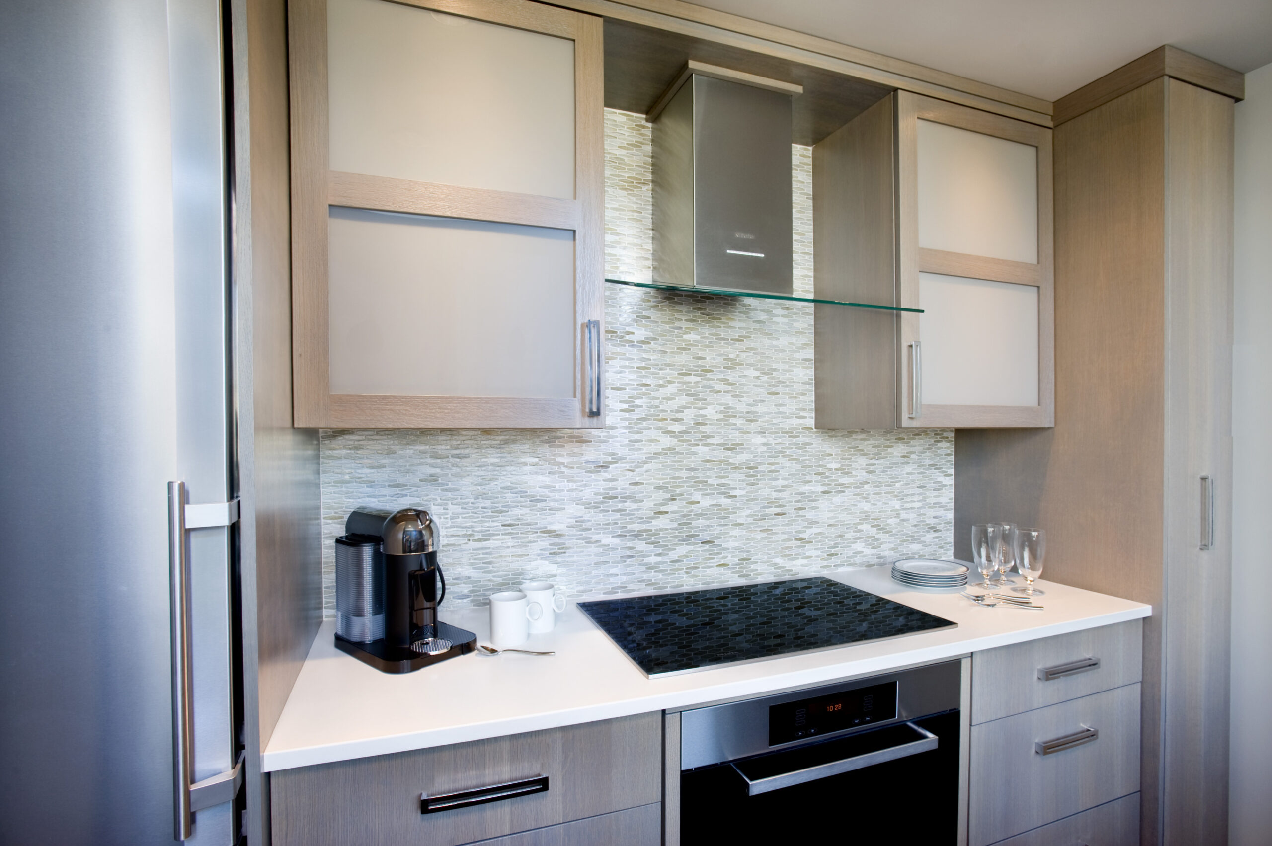 Modern kitchen with light wood cabinets, glass cooktop, built-in oven, coffee machine, and cups set against a mosaic tile backsplash—an elegant example of Luxury Interior Design.