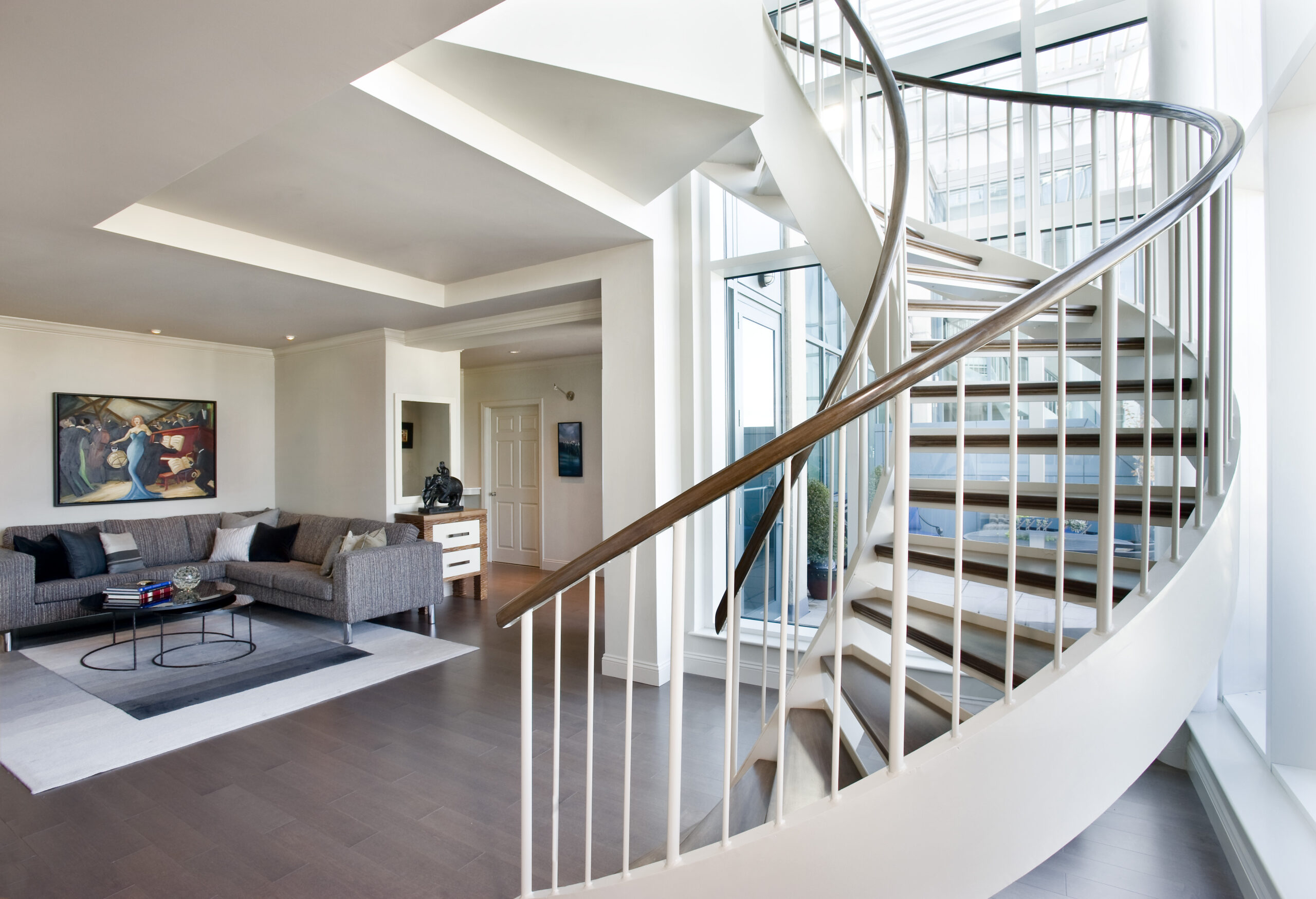 Modern living room with grey couches, a glass coffee table, and a large spiral staircase with white railings and wooden steps; natural light fills this Luxury Interior Design by an expert Interior Designer Massachusetts.