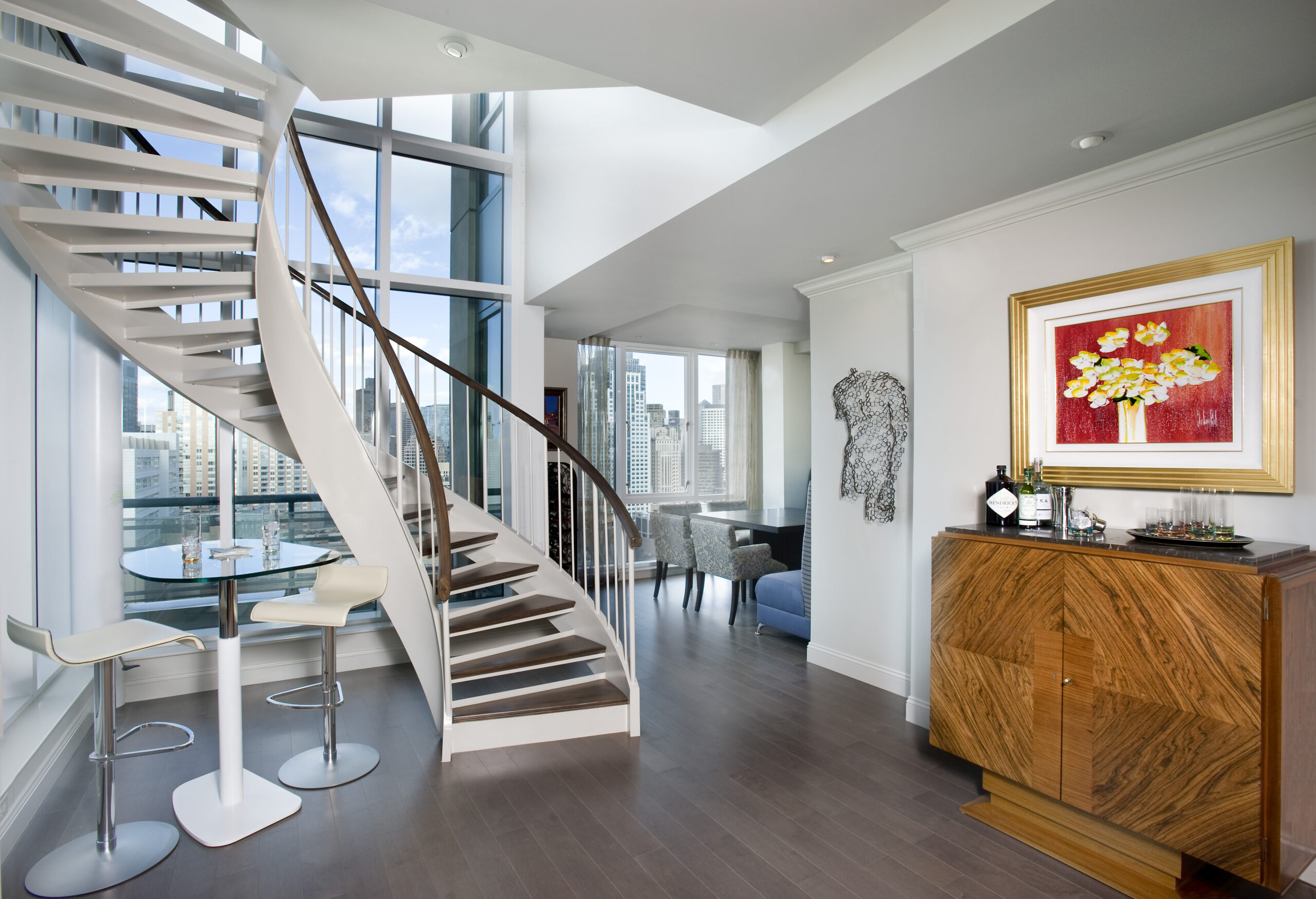 Modern apartment interior with a spiral staircase, large floor-to-ceiling windows, city skyline view, small dining area, wooden cabinet, and wall art—showcasing luxury interior design by a top Interior Designer in Massachusetts.