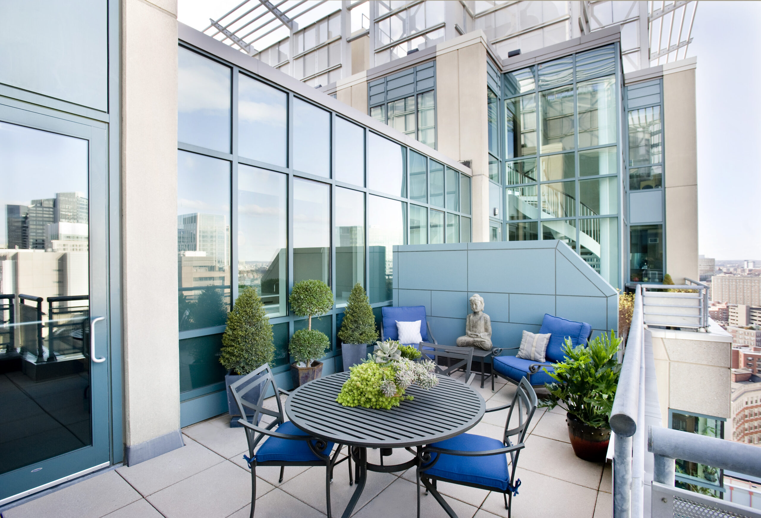 Modern rooftop patio with round metal table, blue cushioned chairs, potted plants, and a Buddha statue—an inviting space that reflects luxury interior design. Surrounded by glass walls, it offers stunning cityscape views.