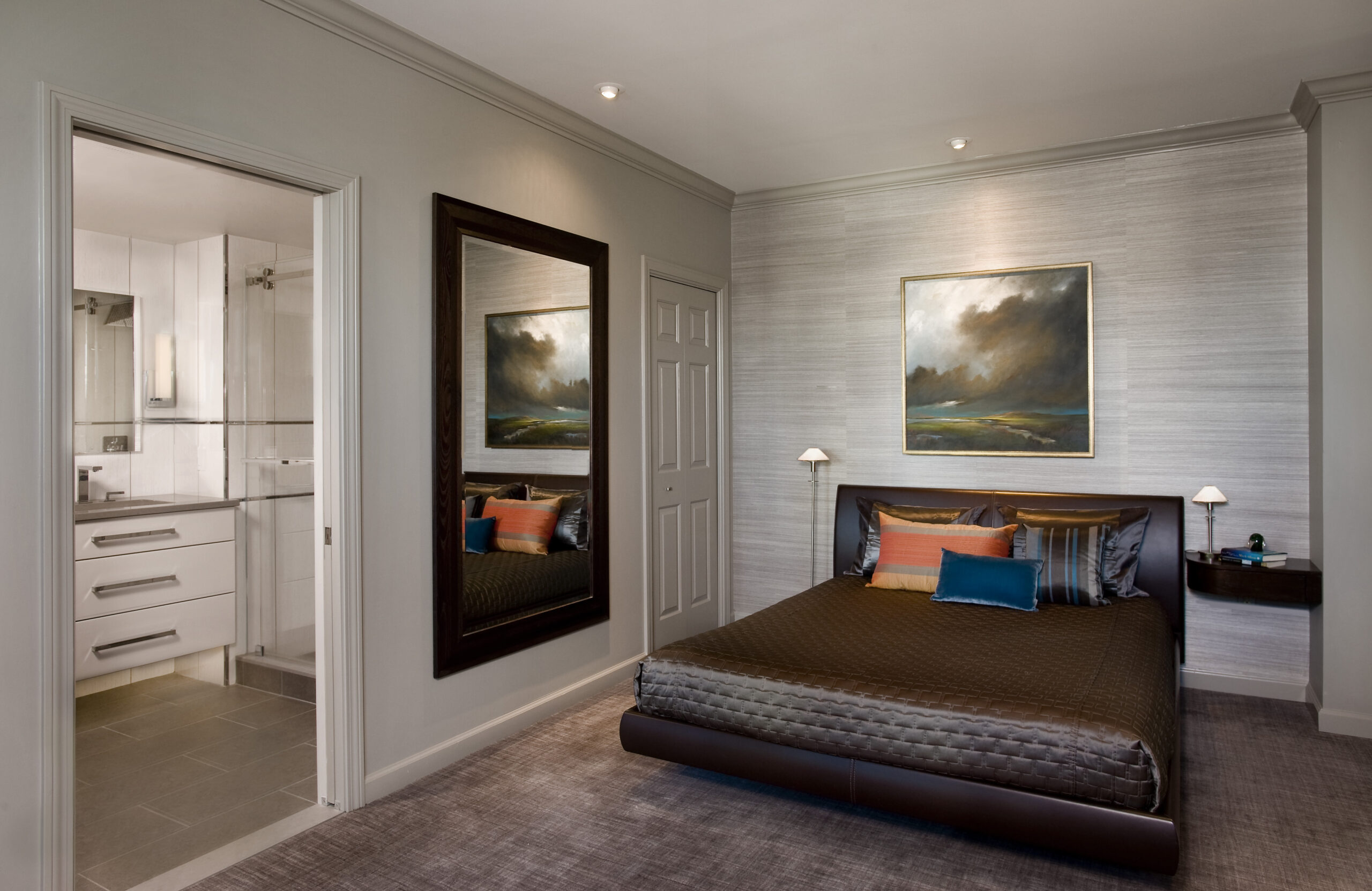 Modern bedroom with a brown bed, colorful pillows, wall art above the headboard, a large wall mirror, and an open door to a white-tiled bathroom—perfectly styled for those seeking luxury interior design in Massachusetts.