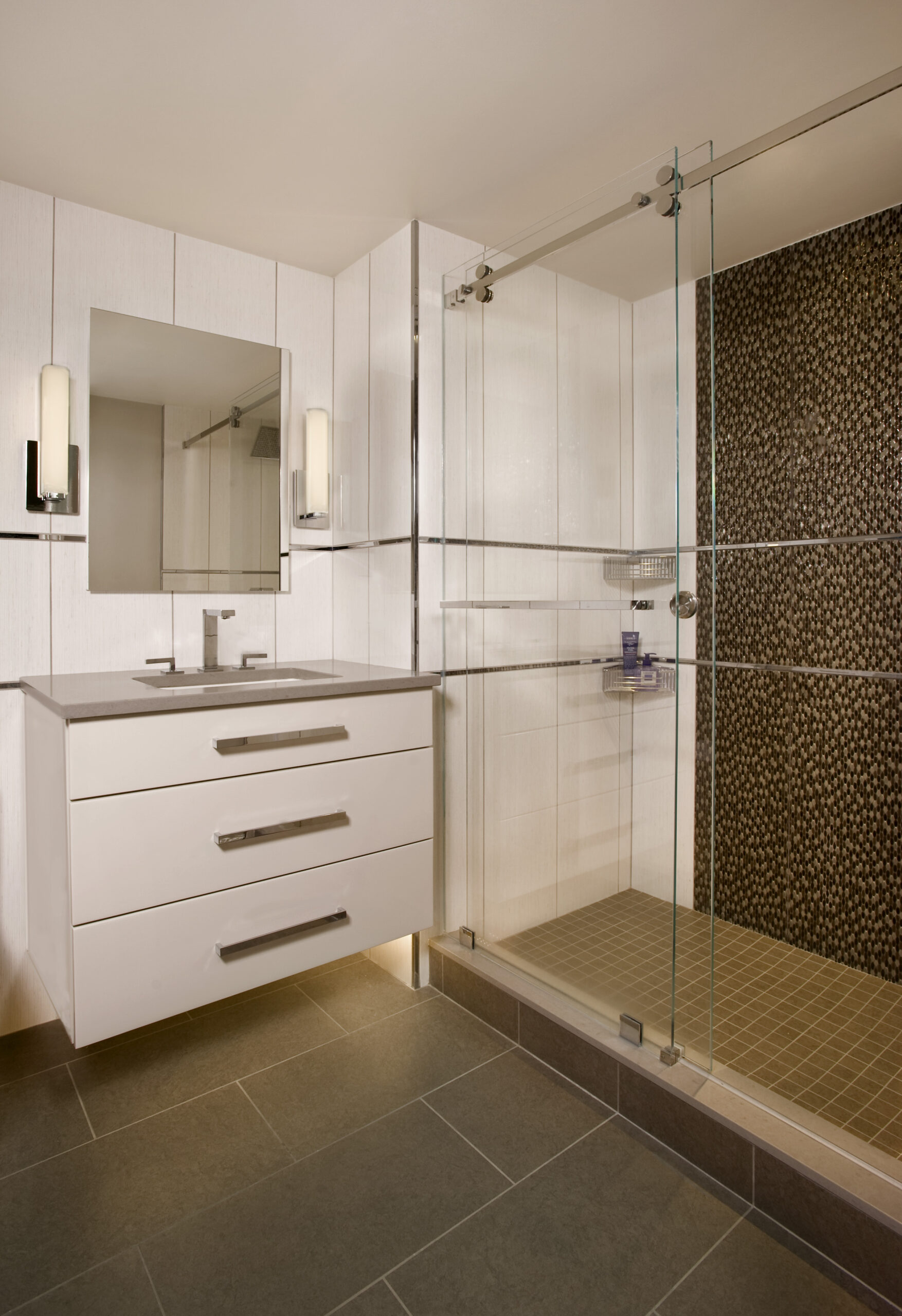 Modern bathroom with a white vanity, rectangular sink, wall-mounted mirror, and a glass-enclosed shower featuring a dark pebble-patterned accent wall—an example of luxury interior design by an interior designer in Massachusetts.