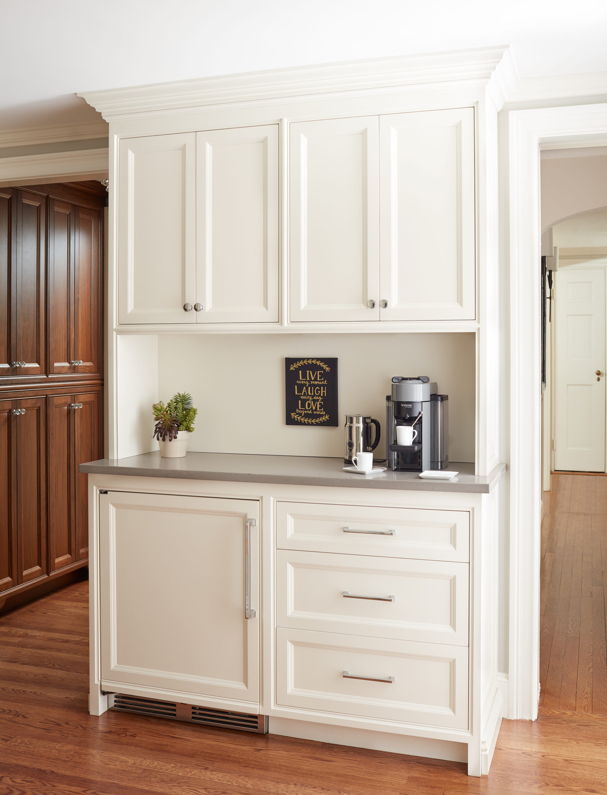 White kitchen cabinets with silver handles, a gray countertop holding a coffee machine, mug, kettle, small plant, and framed sign on the wall. Wood floor and brown cabinets nearby create a touch of luxury interior design.