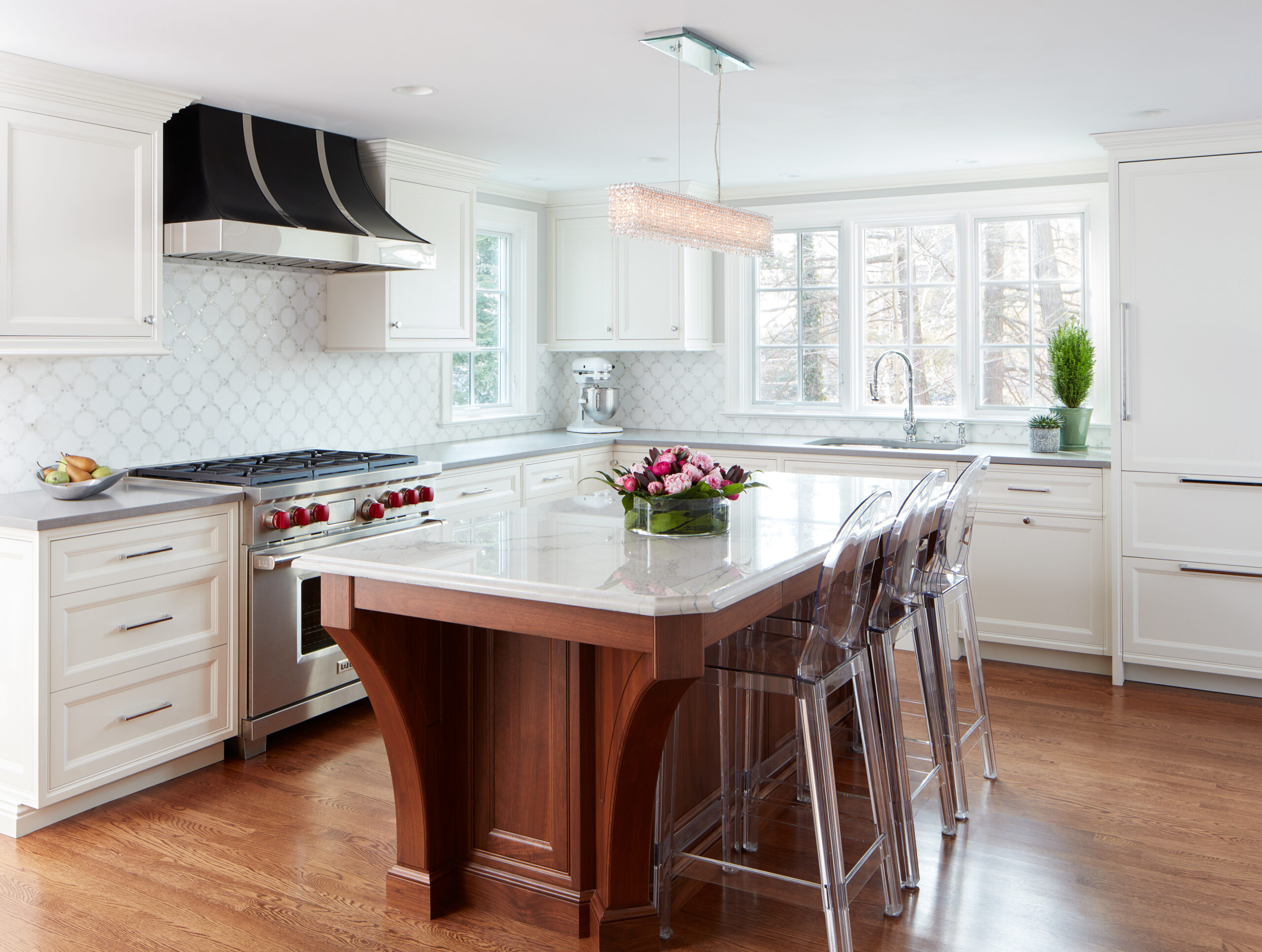 Modern white kitchen with wood island, clear bar stools, stainless steel appliances, and marble countertops. Designed by an expert interior designer in Massachusetts, this luxury interior design features large windows flooding the space with natural light.