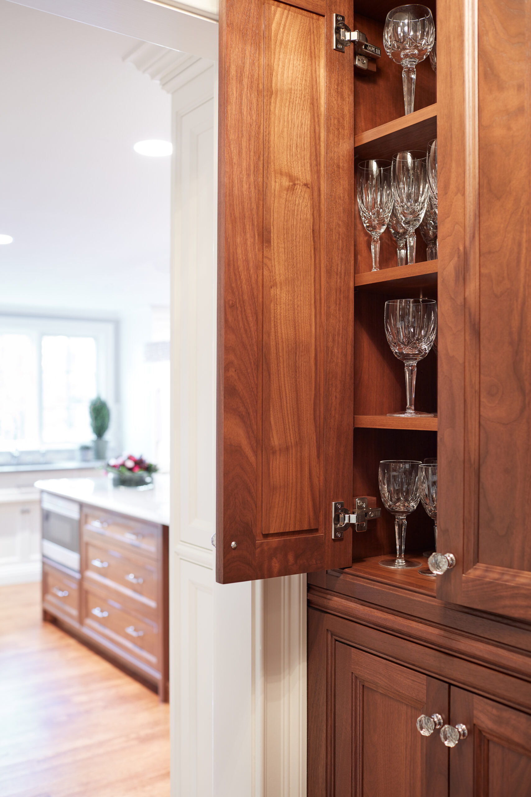 A wooden cabinet with an open door reveals shelves of glassware in a bright kitchen, featuring an island and hardwood floors—a perfect example of luxury interior design.