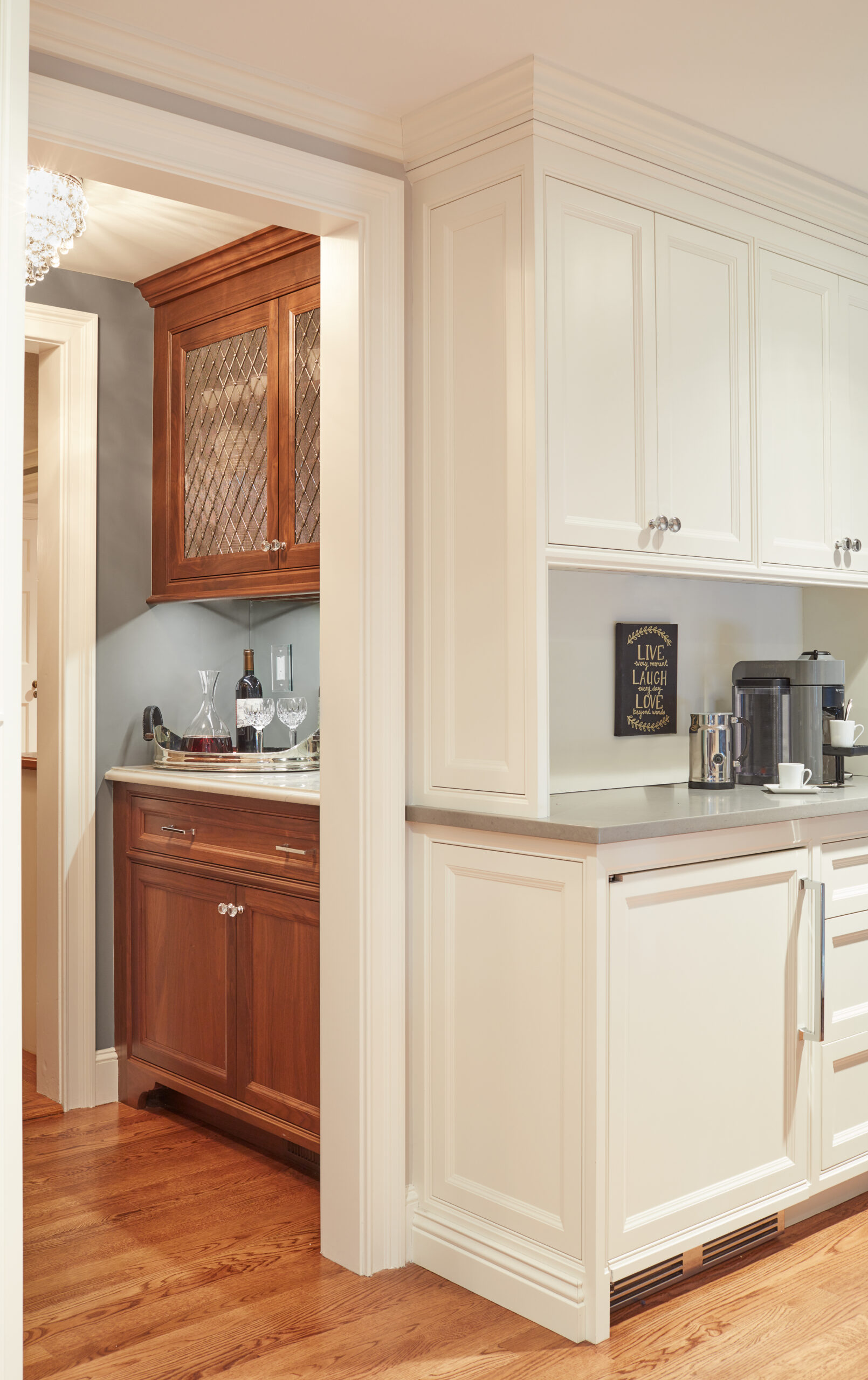 A kitchen with cream-colored cabinets, a coffee maker, and a separate nook featuring wooden cabinetry, wine glasses, and bottles on a counter—crafted with the elegance of luxury interior design by an Interior Designer Massachusetts.