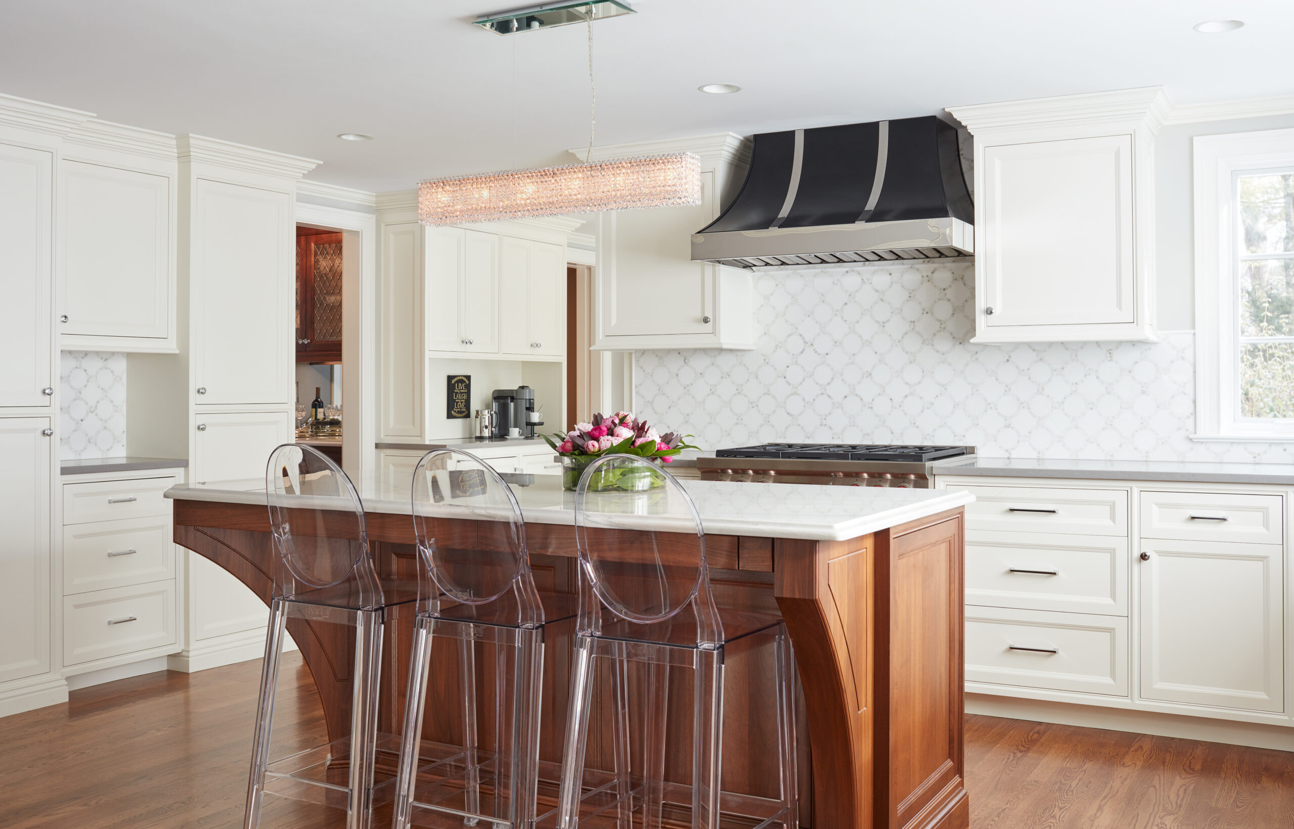 Modern kitchen with white cabinets, a wooden island topped with marble, three clear barstools, a stove with black hood, and a rectangular ceiling light—an example of Luxury Interior Design by an expert Interior Designer Massachusetts.
