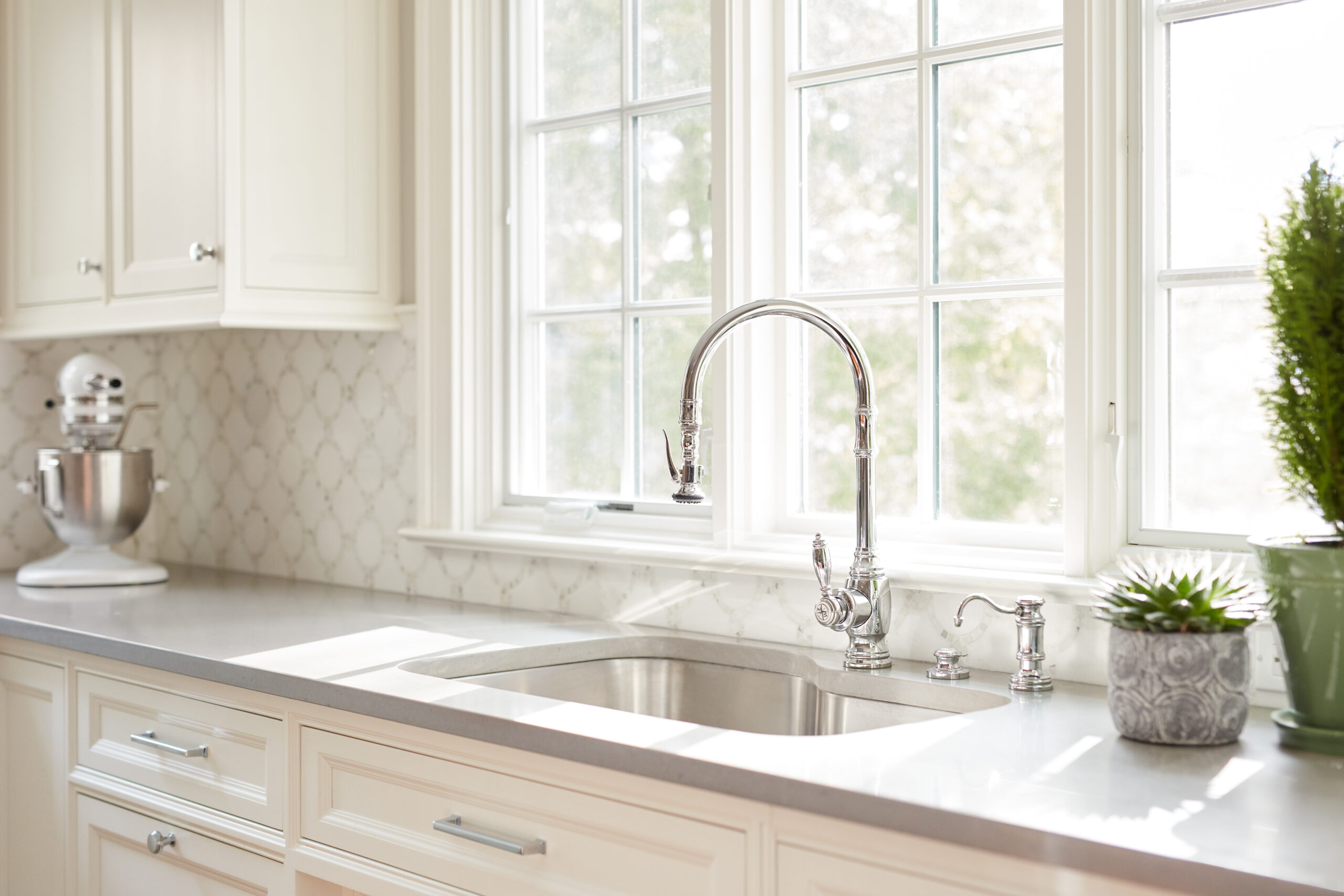 Bright kitchen with a large window, white cabinets, stainless steel sink, silver faucet, potted plants, and a stand mixer on the counter—showcasing touches of luxury interior design.