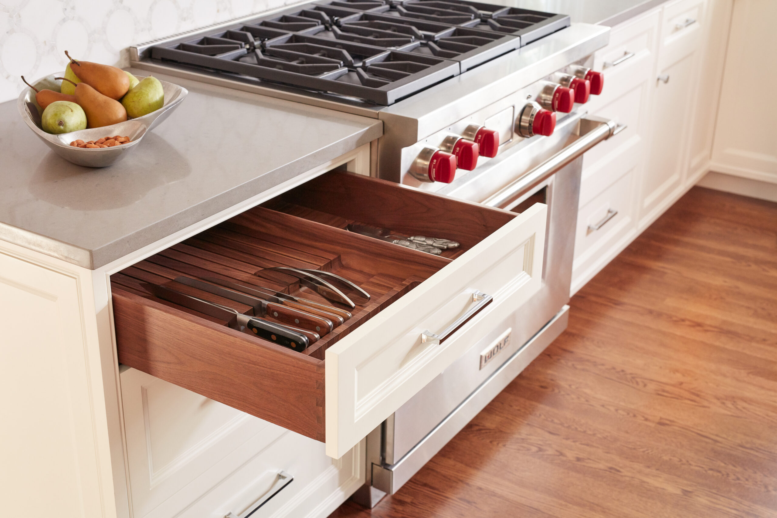 An open kitchen drawer displays organized knives and utensils beside a stainless steel stove with red knobs, reflecting the touch of a Luxury Interior Design specialist. A bowl of fruit sits elegantly on the countertop.