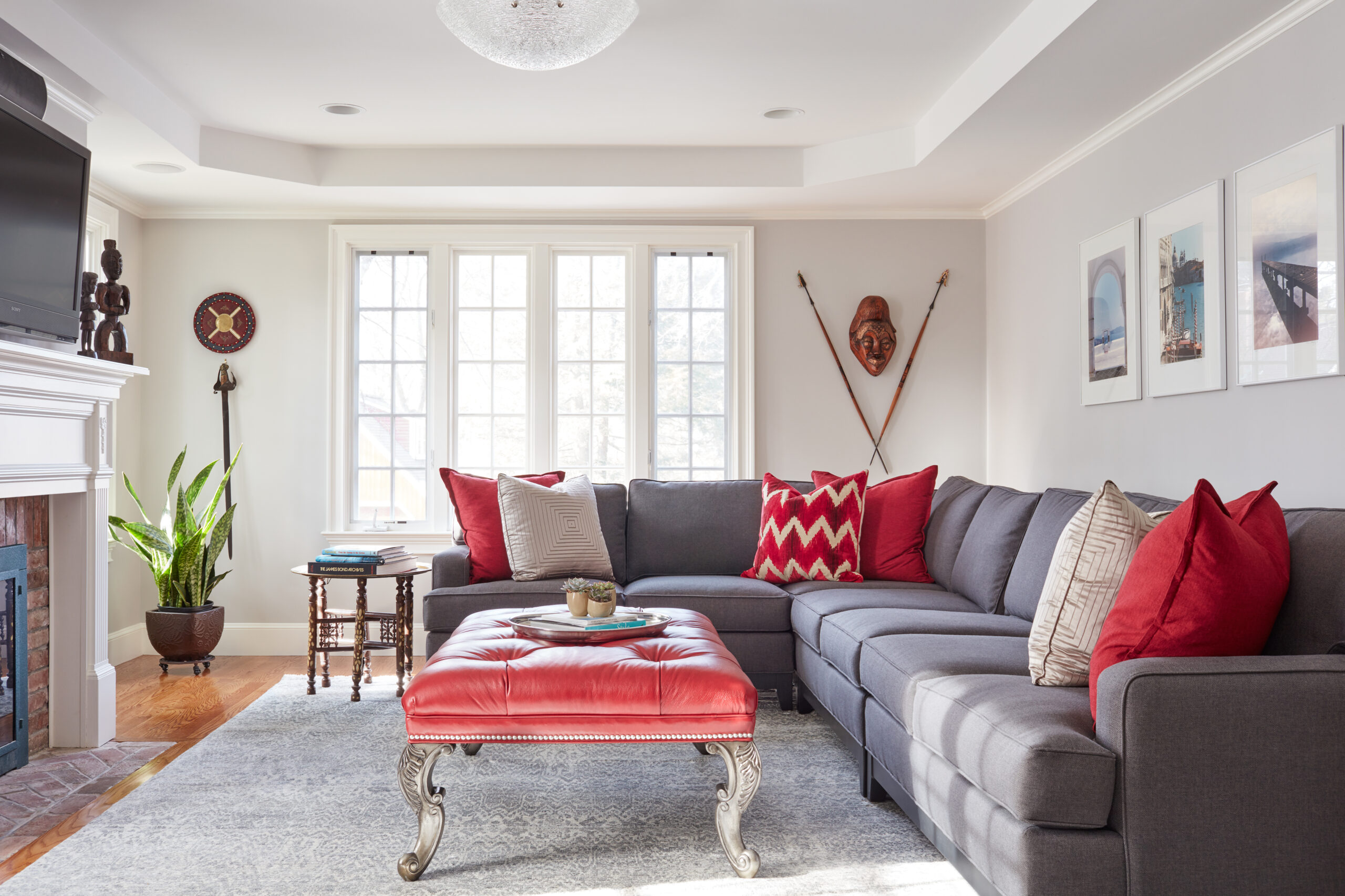 Bright living room with a gray sectional sofa, red and beige accent pillows, a red ottoman, wall art, tribal masks, large windows, and a fireplace with a plant nearby—showcasing luxury interior design by an expert interior designer in Massachusetts.