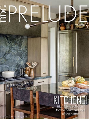 Cover of BDG Prelude magazine featuring a modern kitchen with stone backsplash, built-in appliances, dark countertop, and two wooden chairs; produce and kitchenware on the counter showcase luxury interior design.