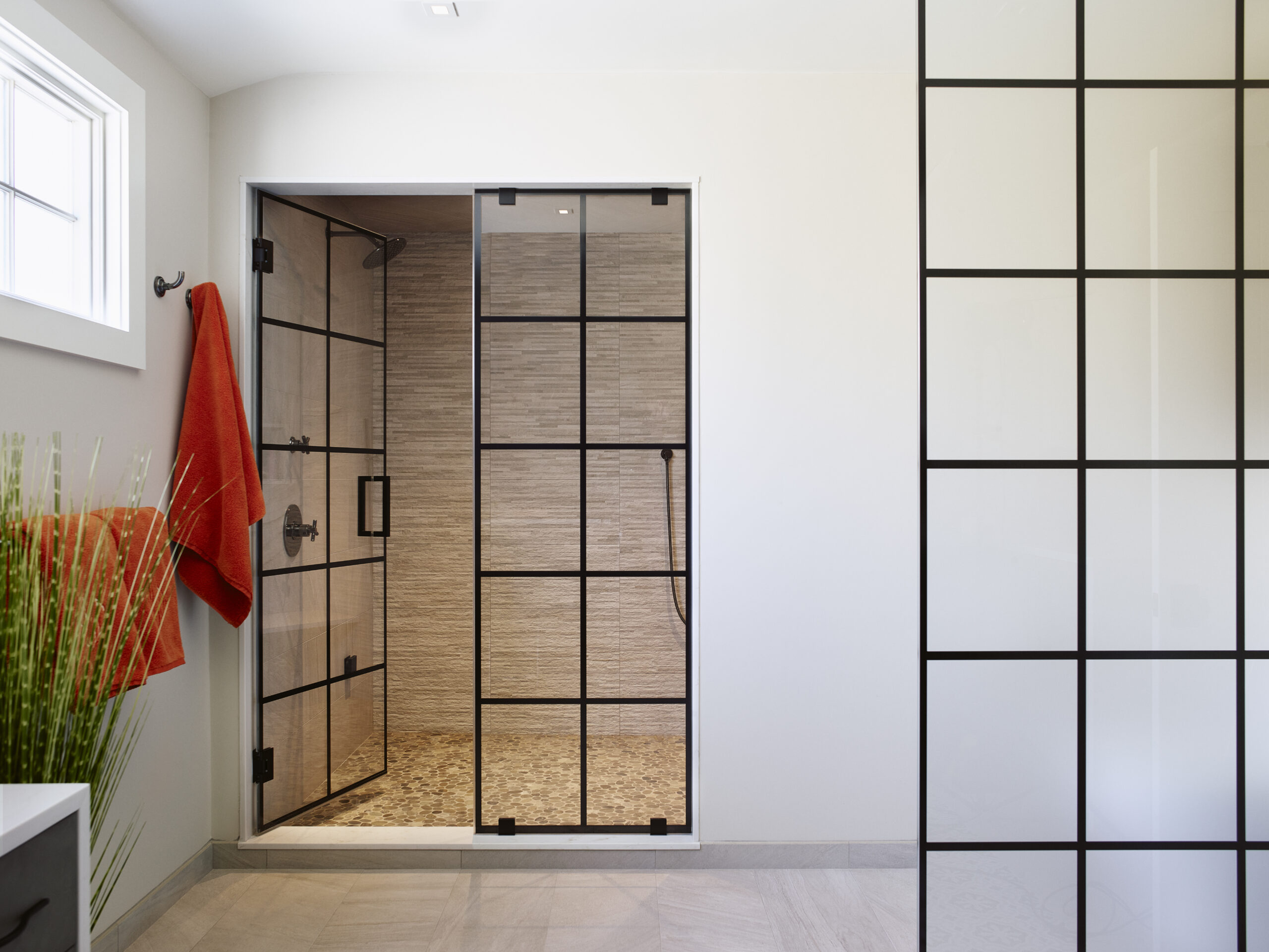 Modern bathroom with a glass shower enclosure featuring black grid frames, a red towel on the wall, and a decorative green plant—an example of luxury interior design curated by an expert interior designer in Massachusetts.