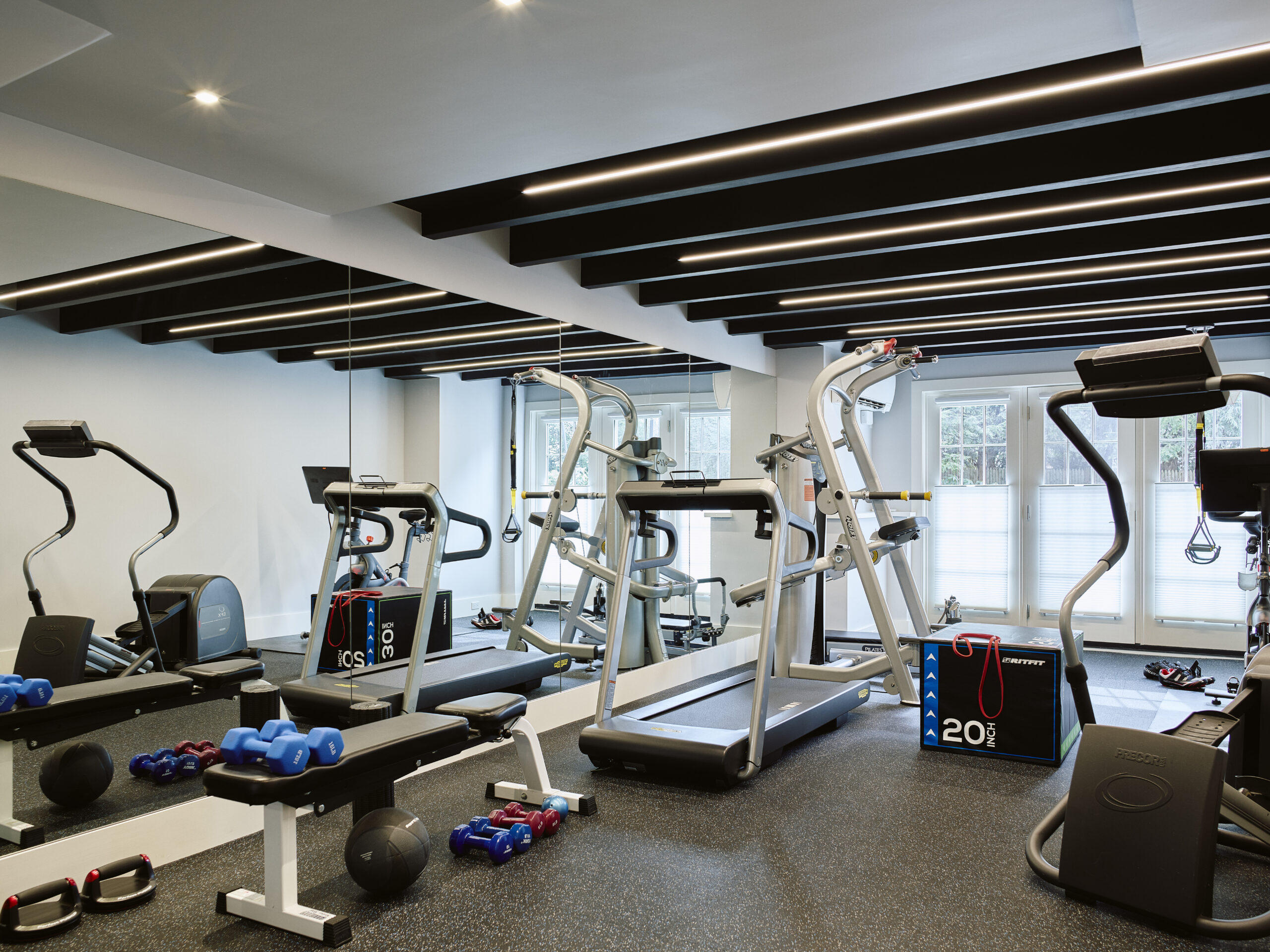Modern gym with treadmills, elliptical machines, weight benches, dumbbells, kettlebells, and resistance bands. Designed by a top Interior Designer in Massachusetts for a luxury interior design feel. Large mirrors and natural light complete the space.