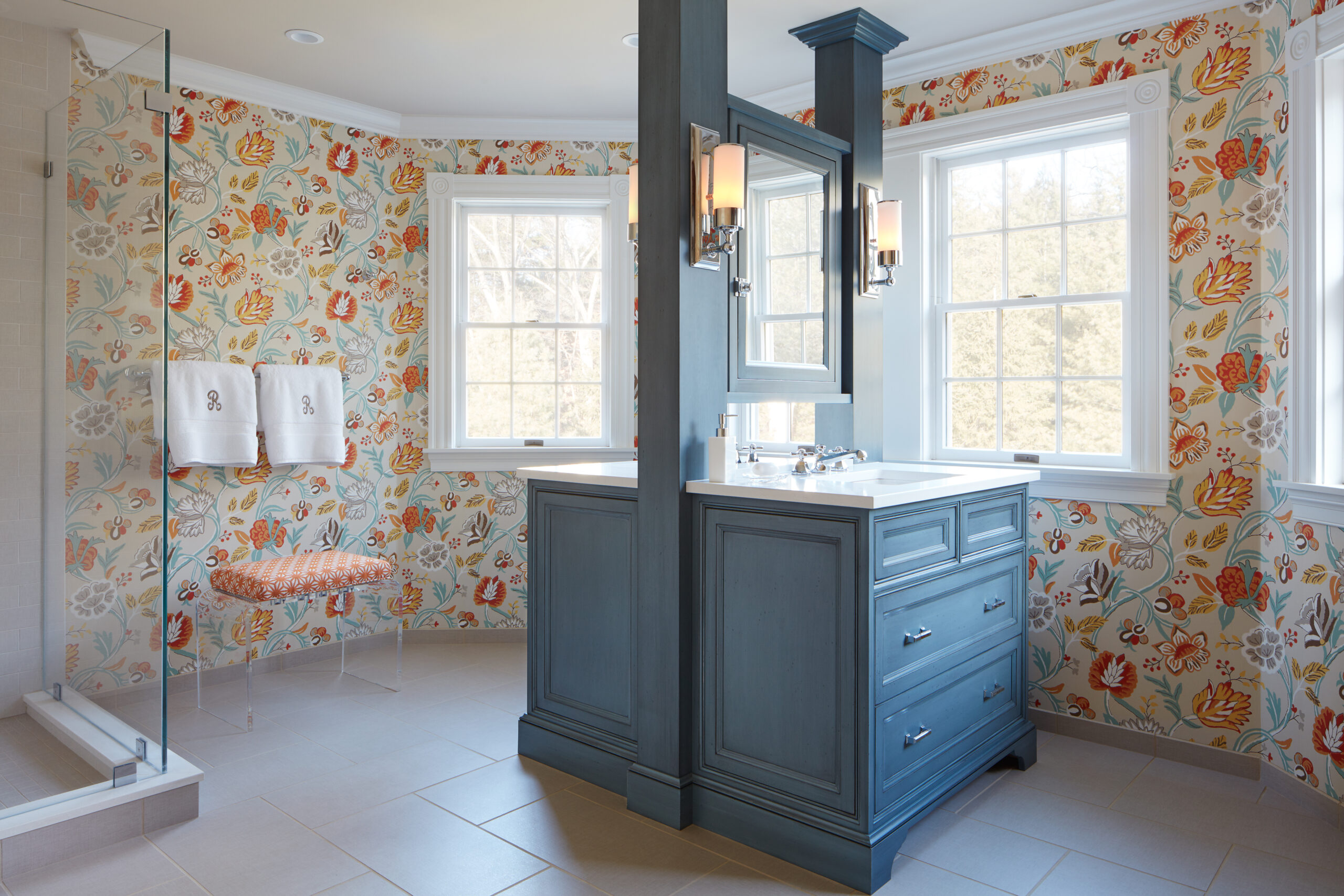 This luxury interior design bathroom features floral wallpaper, a blue double-sink vanity, two windows, a glass shower, a bench with an orange cushion, and two folded towels—perfect inspiration from an Interior Designer Massachusetts.