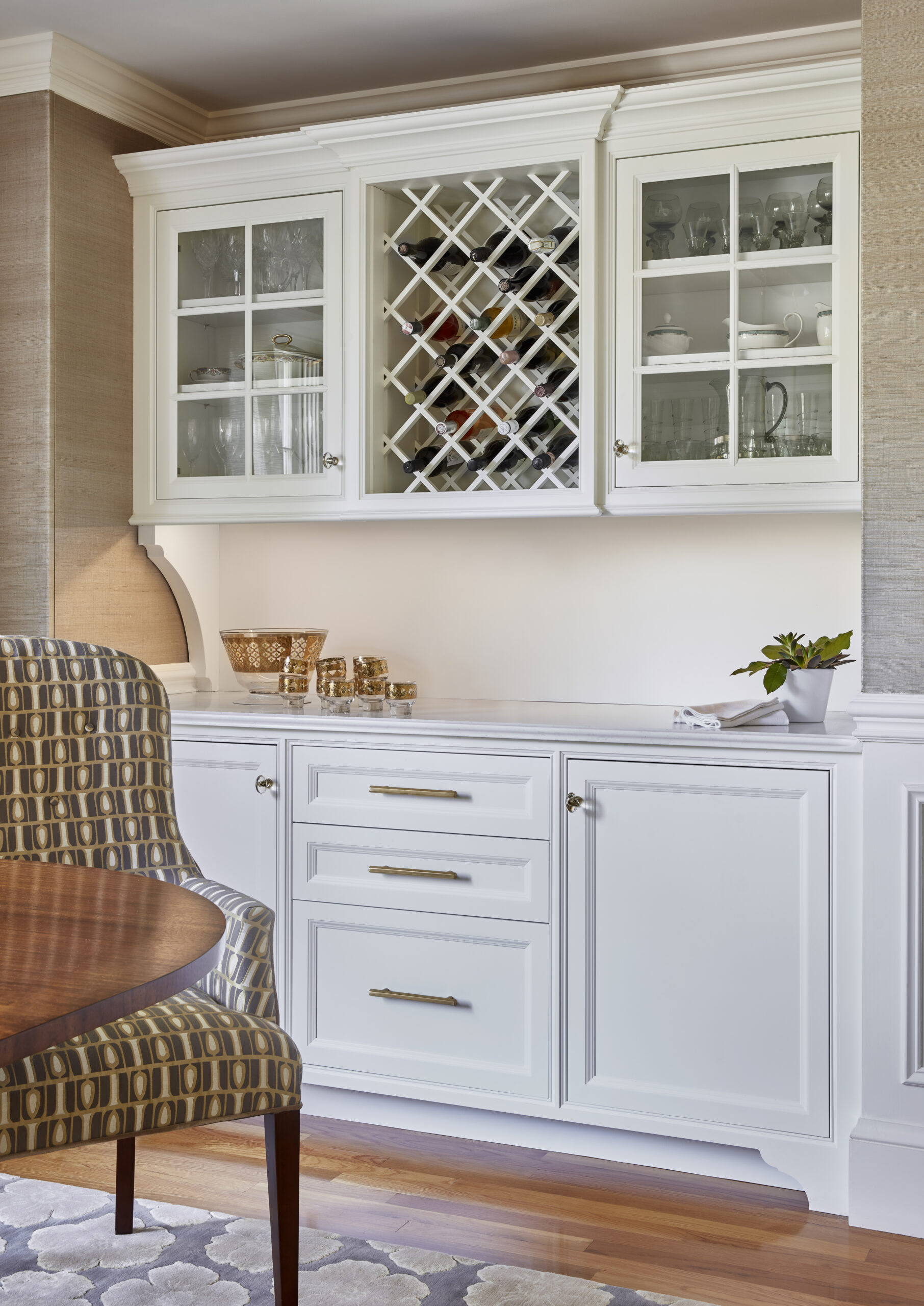 White built-in cabinetry with glass-front upper cabinets, a wine rack, and countertop, curated by a luxury interior design expert, sits next to a patterned upholstered chair and round wooden table.