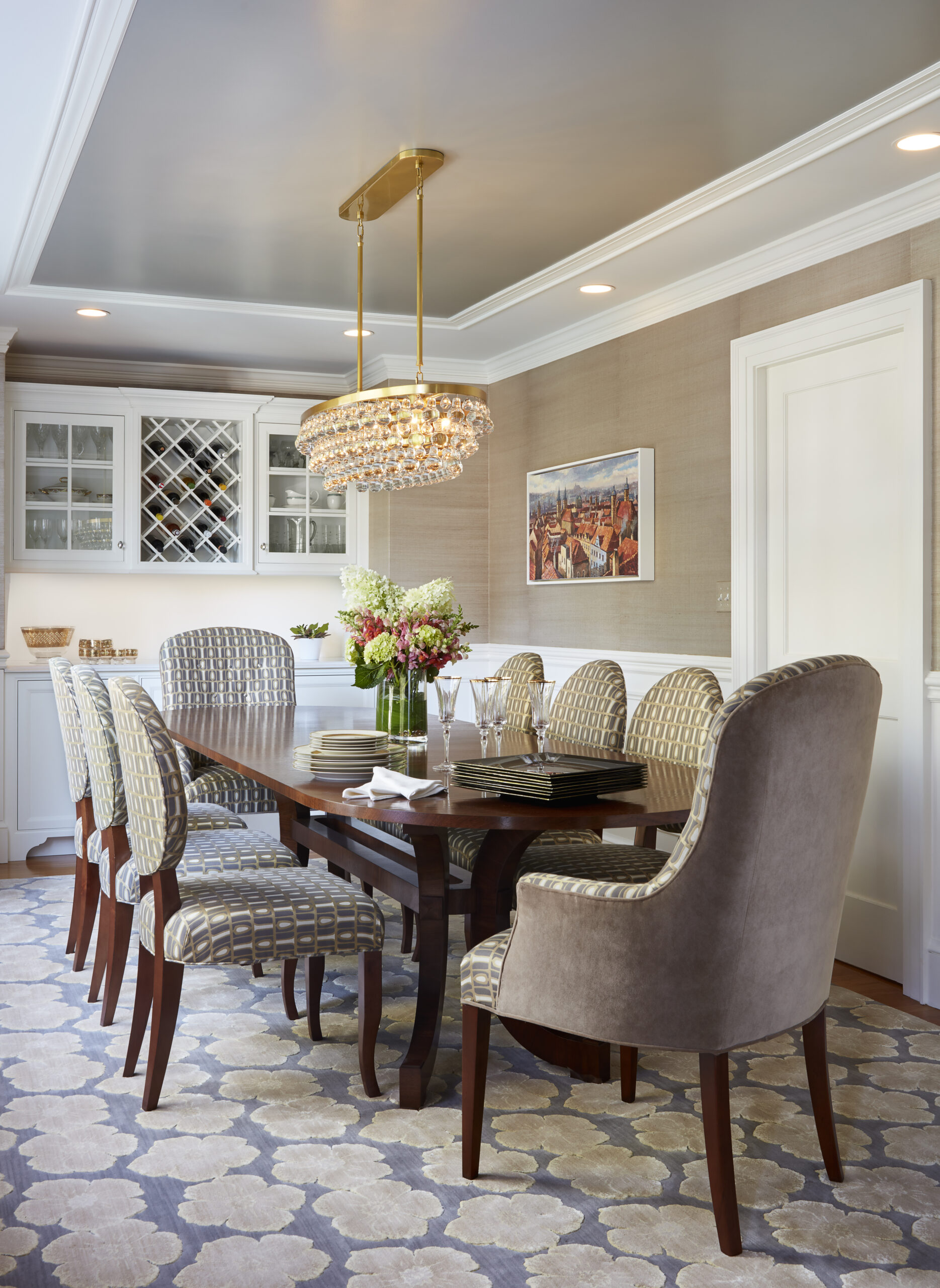A contemporary dining room by an Interior Designer Massachusetts features a wooden table, eight patterned upholstered chairs, a floral centerpiece, chandelier, cabinet with wine storage, and elegant framed wall art for a touch of luxury interior design.