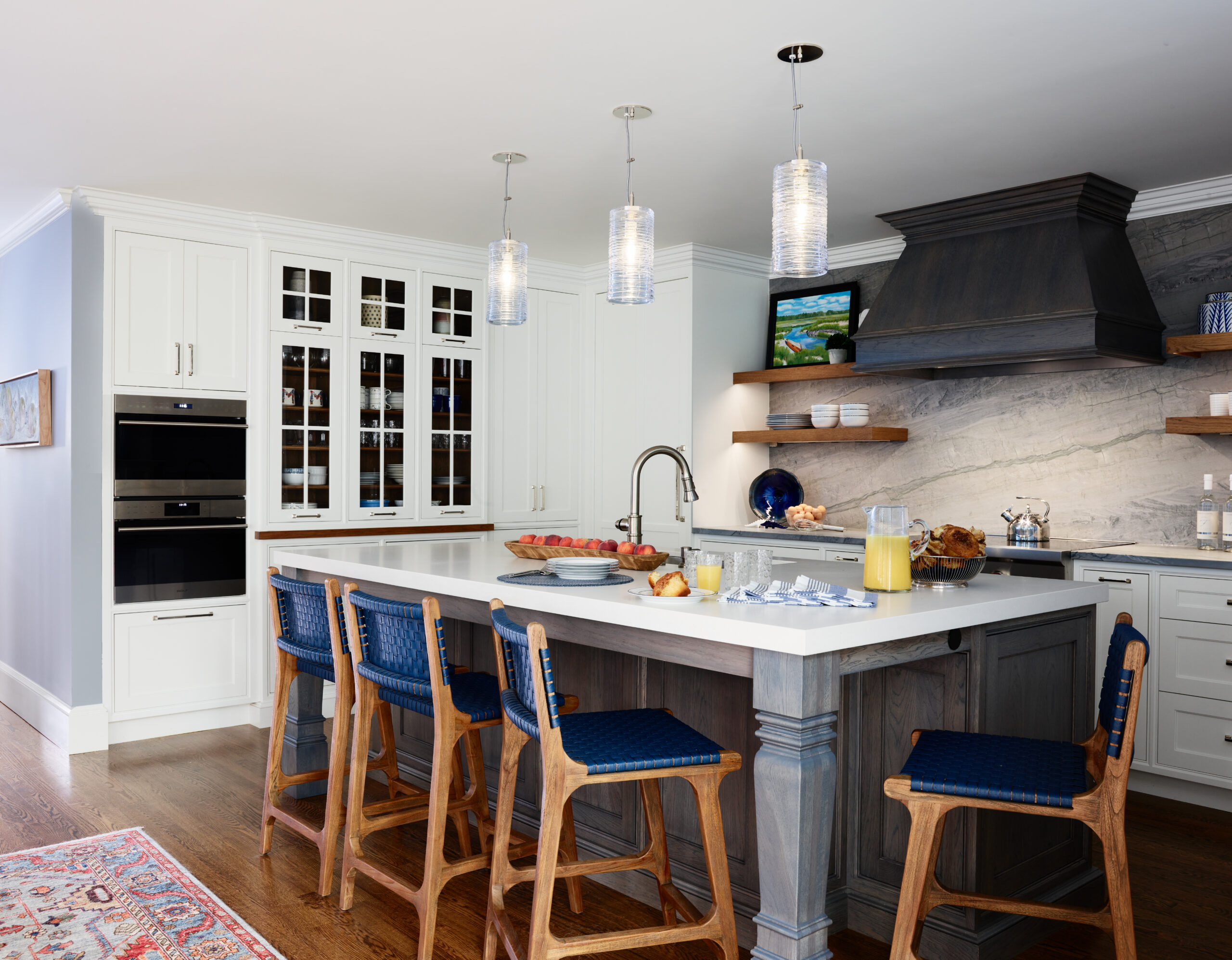 Modern kitchen with a large island, blue cushioned bar stools, pendant lights, built-in ovens, open shelves, marble backsplash, and wooden flooring—showcasing luxury interior design by an expert Interior Designer Massachusetts.