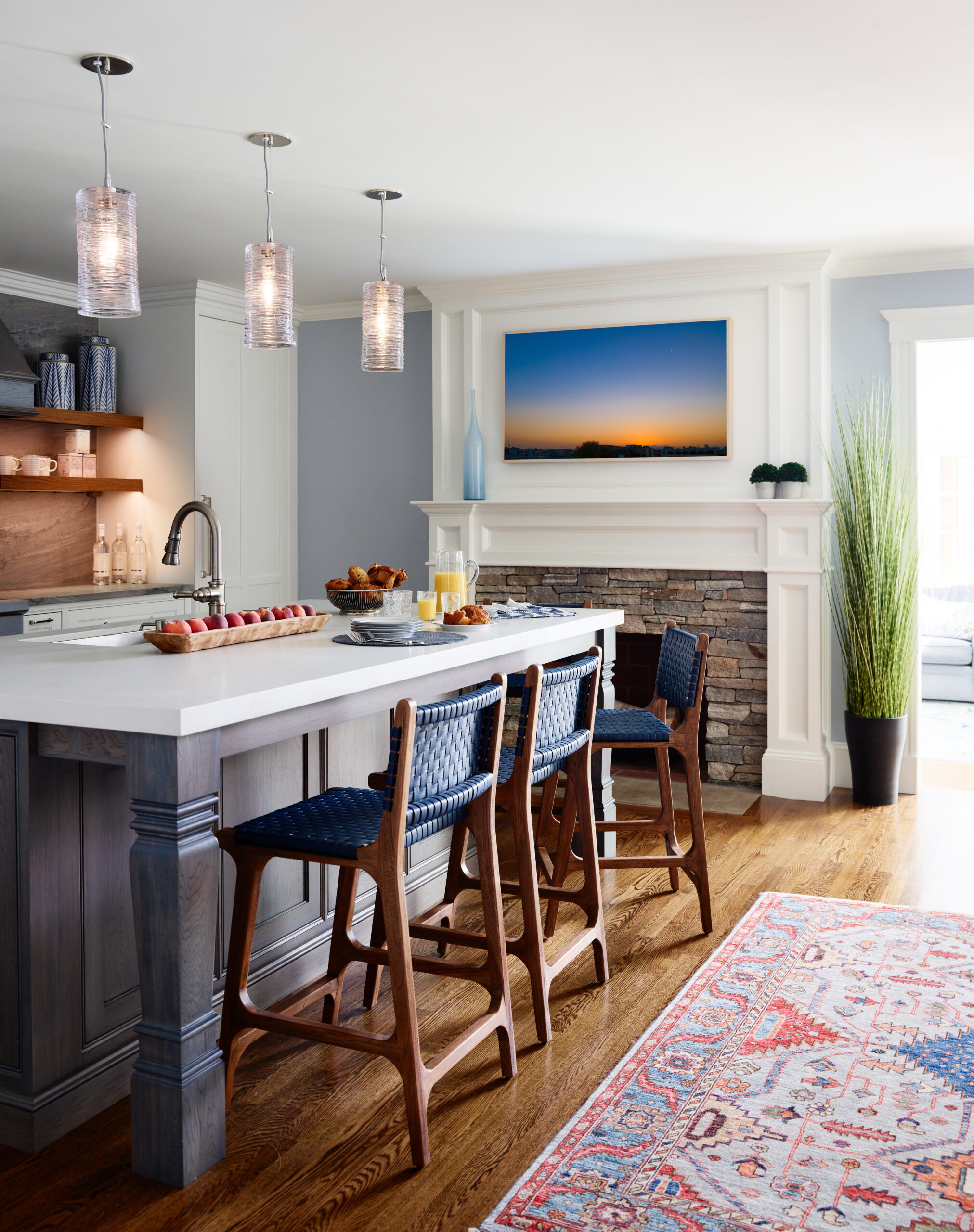 Modern kitchen with a large island, three blue woven barstools, pendant lights, fireplace with a sunset photo above, and a tall plant by the mantel—an example of Luxury Interior Design by an expert Interior Designer Massachusetts.