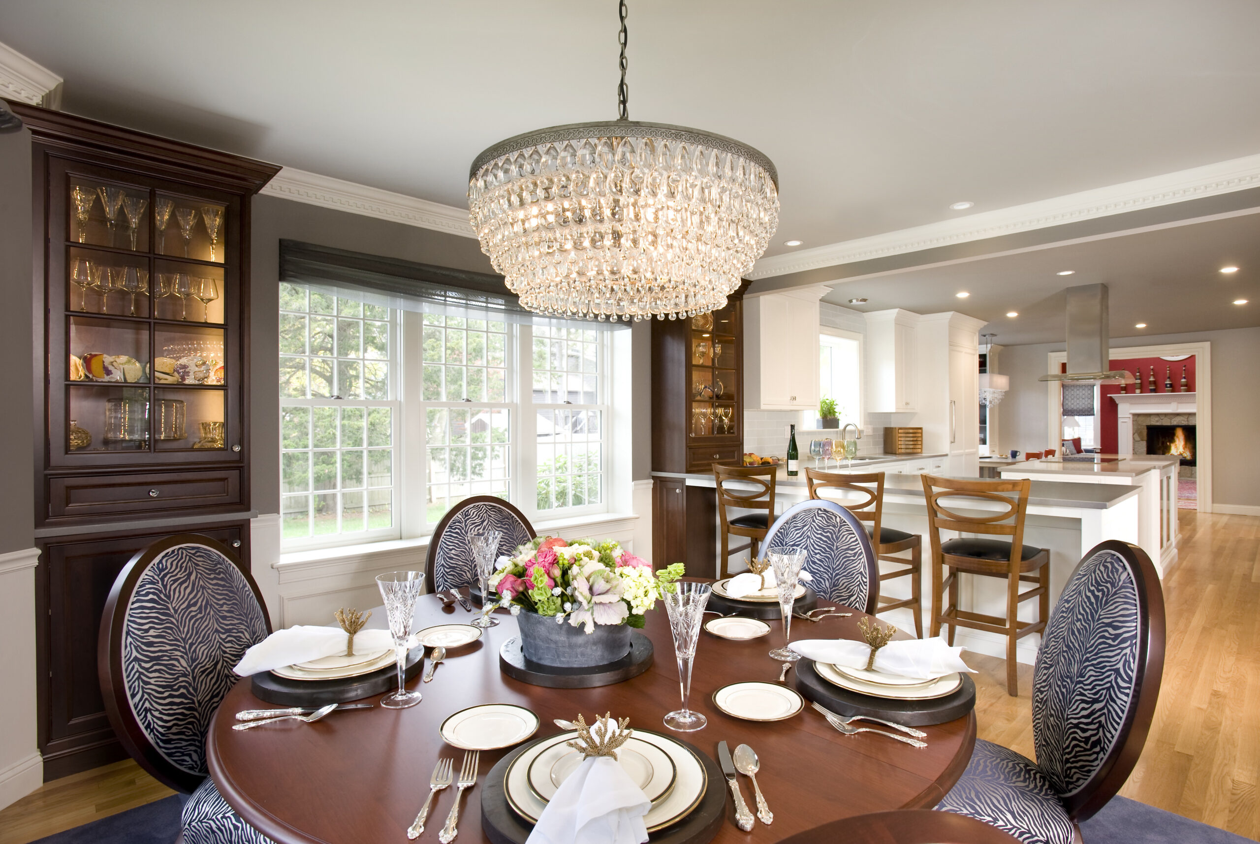 A dining room set for six features a floral centerpiece, crystal chandelier, and an open view into a modern kitchen with barstools and hardwood floors—showcasing luxury interior design by an expert interior designer in Massachusetts.
