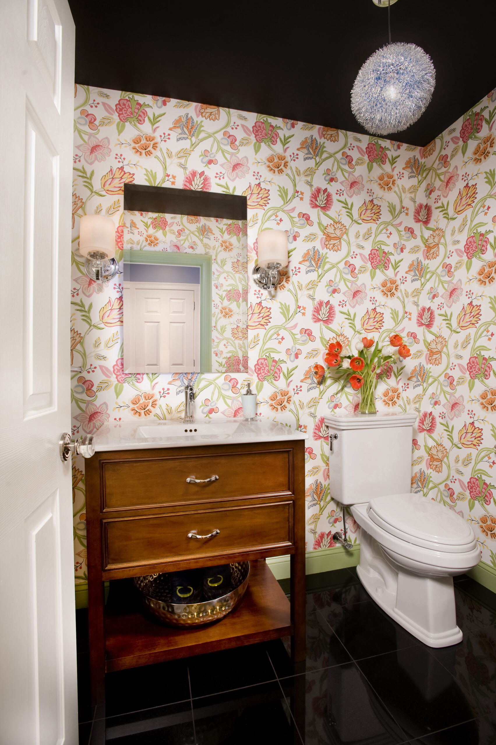 A bathroom with floral wallpaper, a wooden vanity and decorative sink, mirror, toilet, modern lighting, and a vase of flowers on the toilet tank showcases luxury interior design by a talented interior designer in Massachusetts.