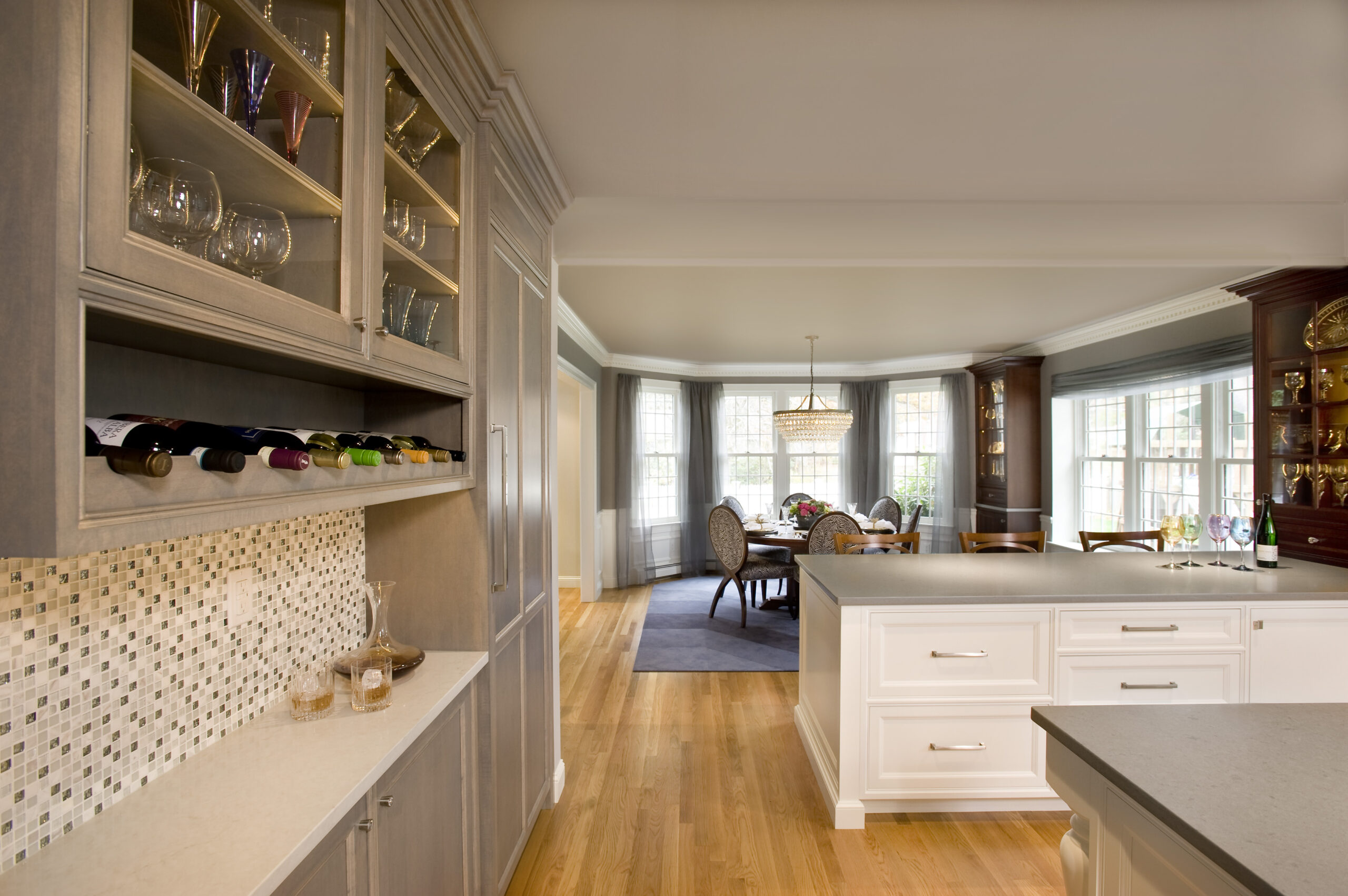 A modern kitchen with light wood floors, white cabinets, a built-in wine rack, and glassware displays showcases luxury interior design, offering a stunning view into a dining area with large windows.