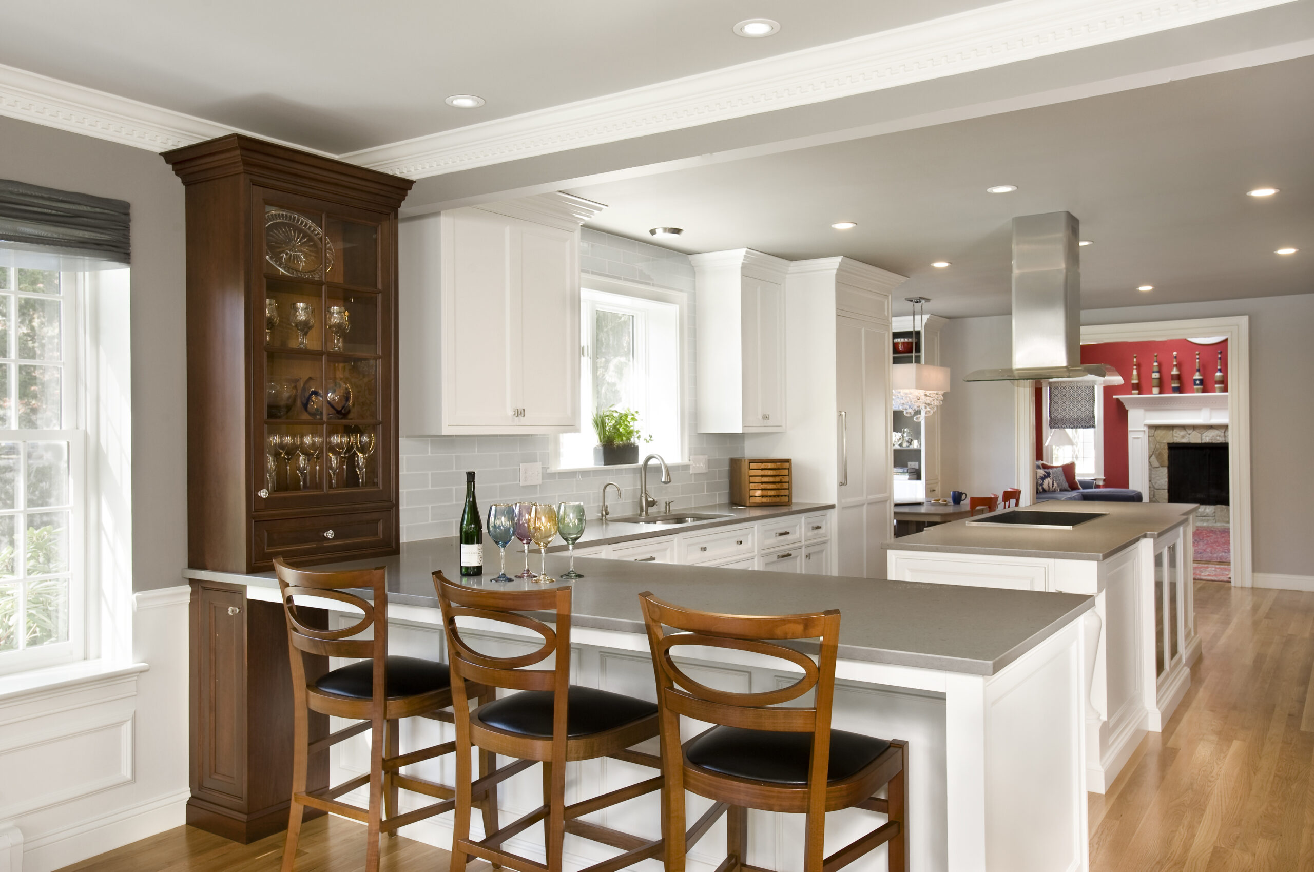 Modern kitchen with white cabinets, a large island with three wooden barstools, stainless steel appliances, and a glass cabinet displaying glasses—a stunning example of Luxury Interior Design by an Interior Designer Massachusetts.