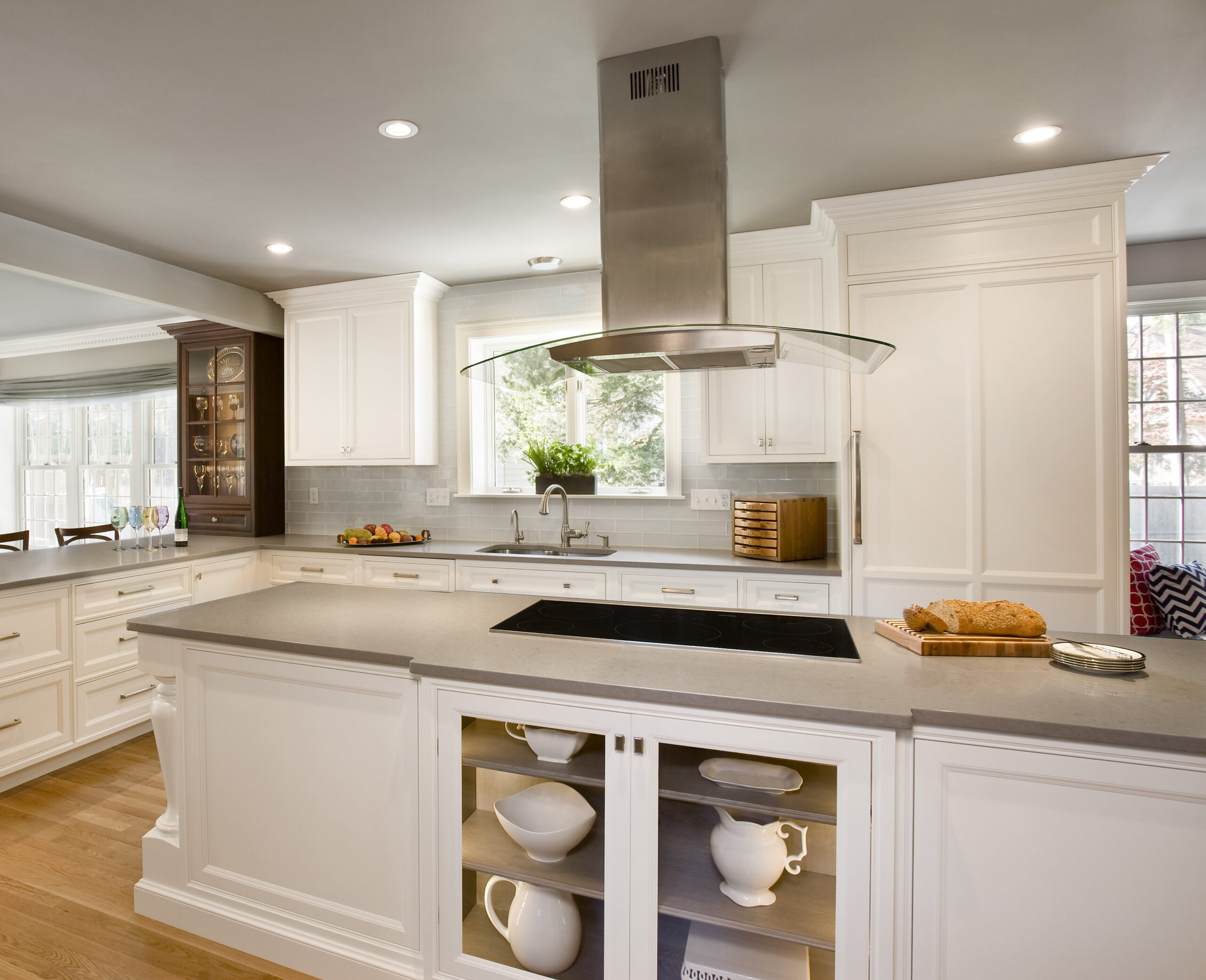 Modern kitchen with white cabinets, a large island featuring an electric cooktop, open shelves displaying dishes, and a stainless steel range hood under recessed ceiling lights—an example of luxury interior design by an Interior Designer Massachusetts.