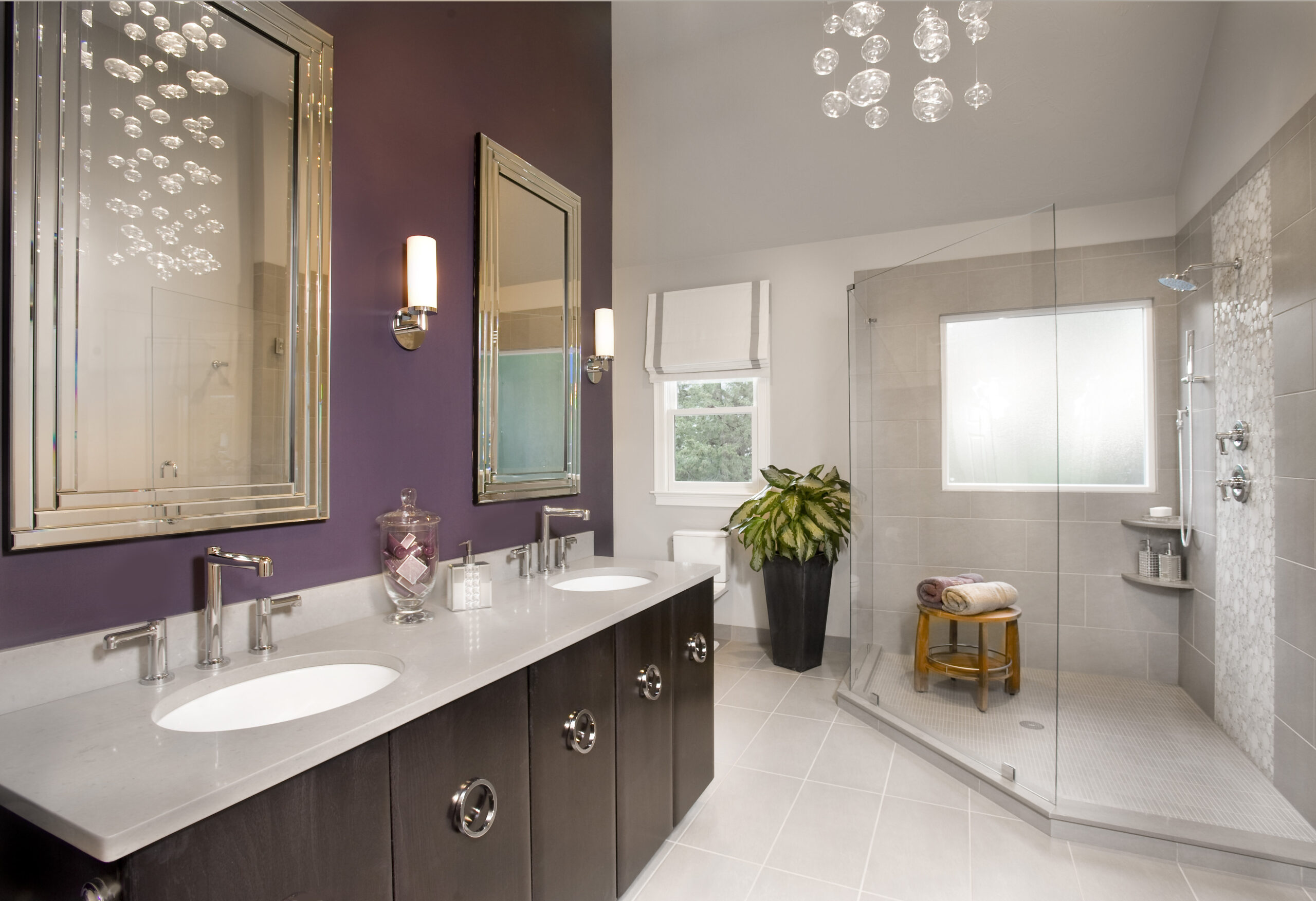 Modern bathroom with double sinks, large mirrors, wall sconces, a glass-enclosed shower, potted plant, and purple accent wall. Luxury Interior Design touches include a towel on a stool and stylish jars on the counter.