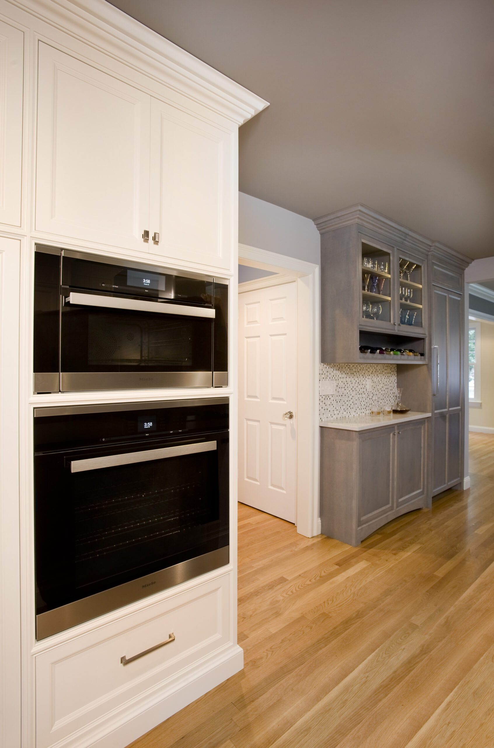 Modern kitchen with built-in double ovens, white cabinetry, light wood flooring, and a gray backsplash area with glass shelves—showcasing luxury interior design by an expert interior designer Massachusetts residents trust.