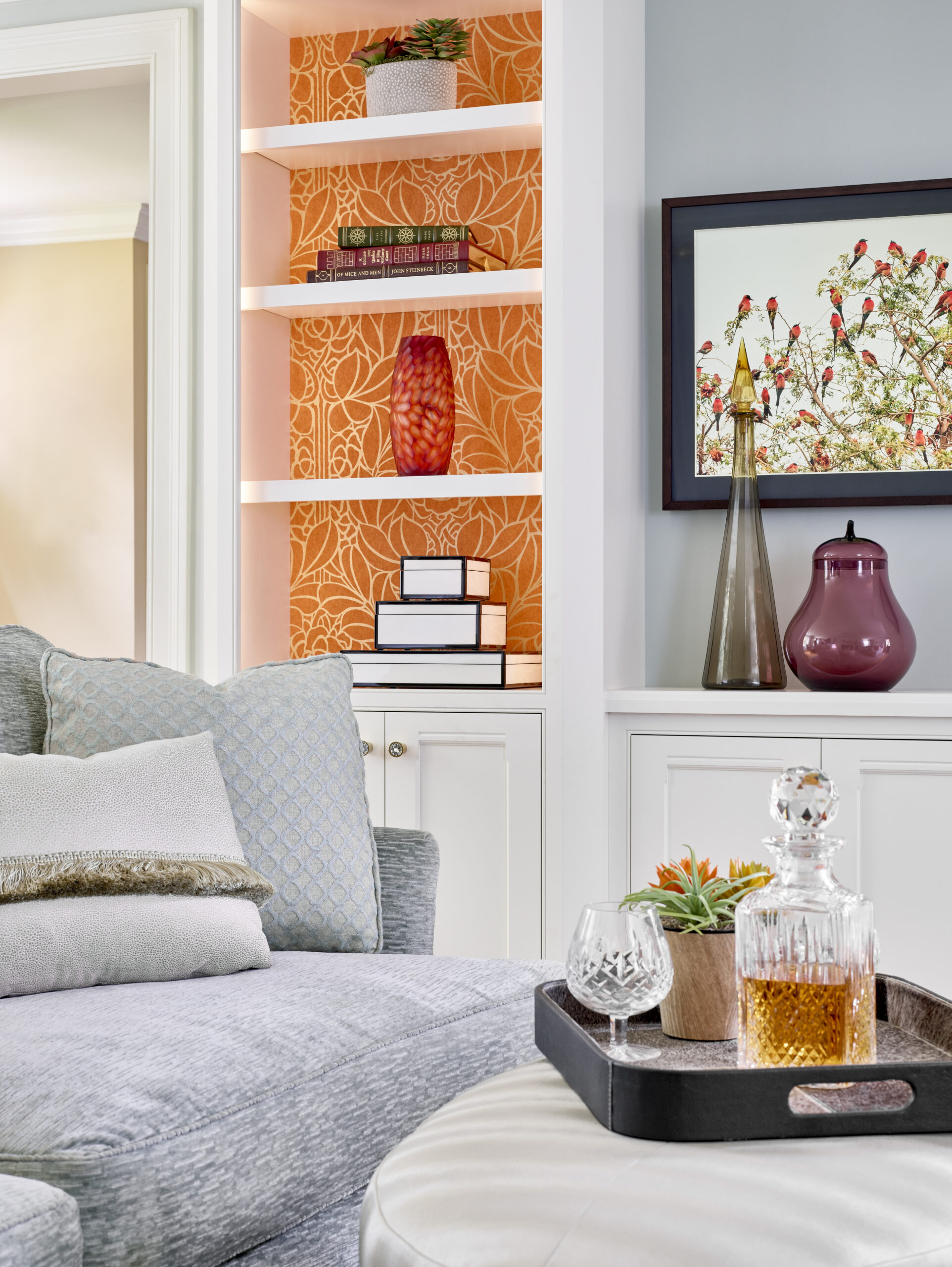 A living room with a gray sofa, built-in shelves with orange wallpaper, decorative objects, and a tray holding a decanter, glasses, and a small plant on an ottoman showcases luxury interior design by an expert interior designer in Massachusetts.
