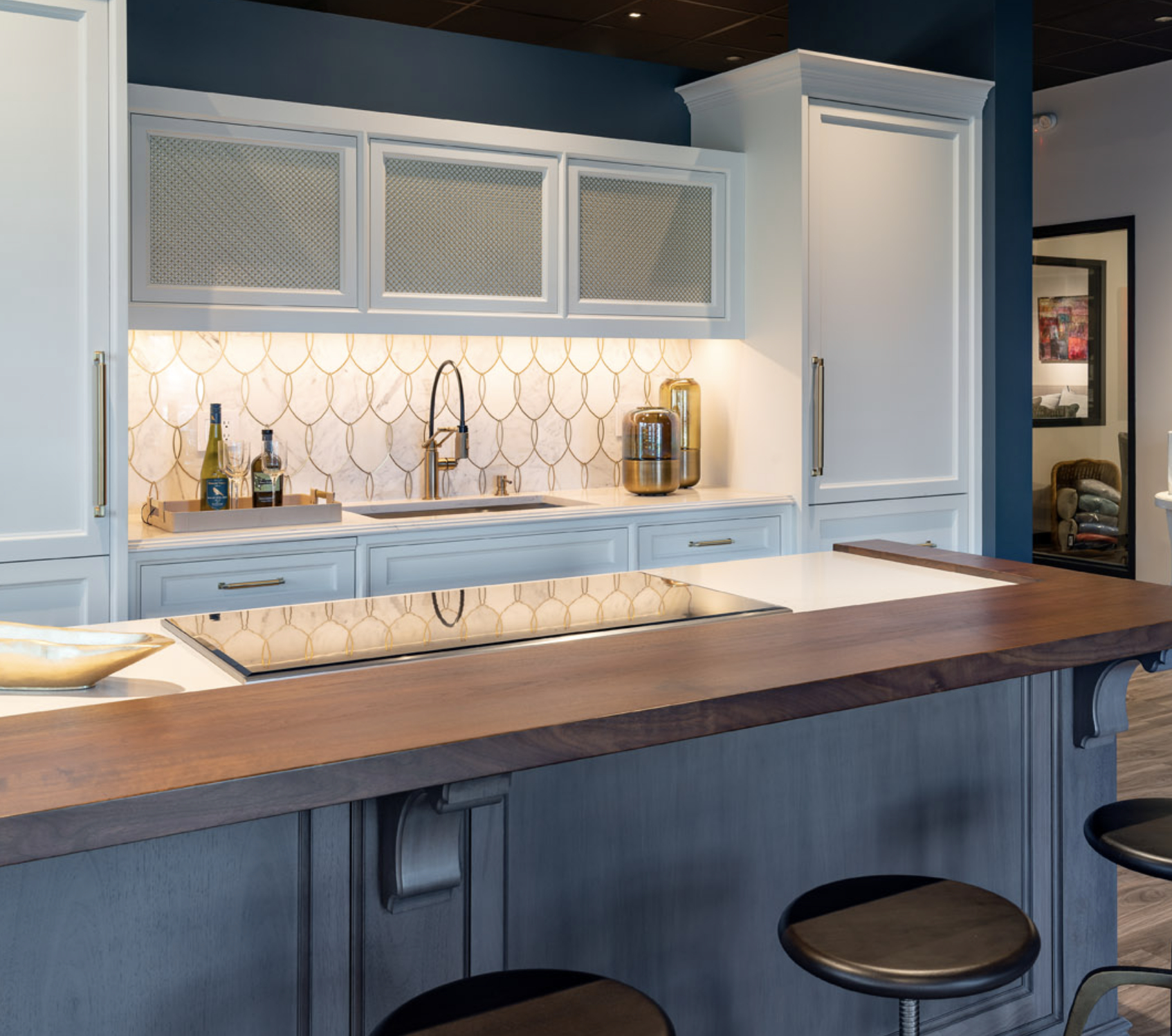 Modern kitchen with white cabinets, patterned tile backsplash, built-in stove, sink, and wooden island with bar stools—showcasing luxury interior design by a top interior designer in Massachusetts. Bottles and appliances line the countertop.