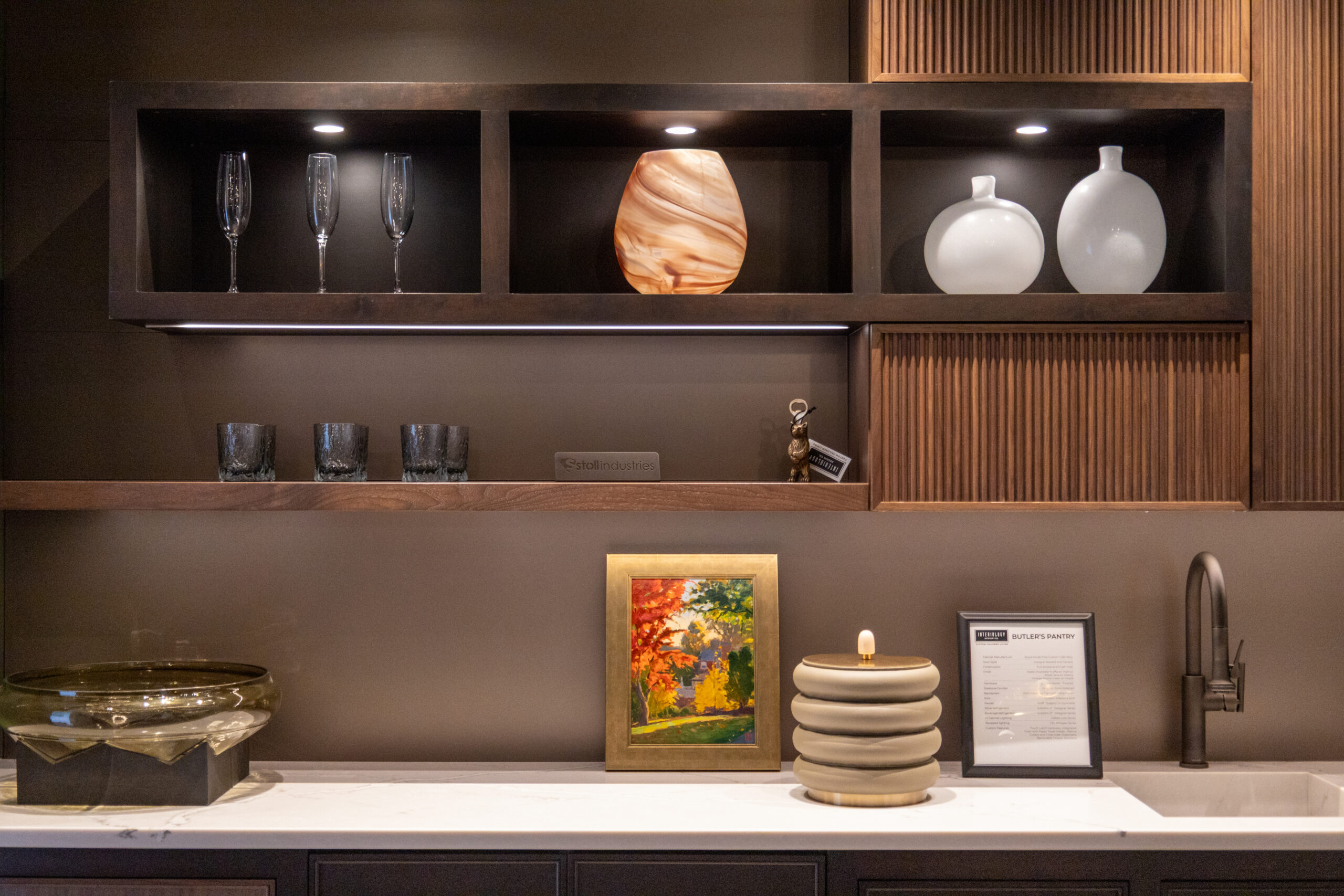 Modern kitchen countertop with decorative items, framed art, and open shelves displaying glassware—crafted with luxury interior design touches—plus two white vases and a marbled round vase, all illuminated by warm lighting.