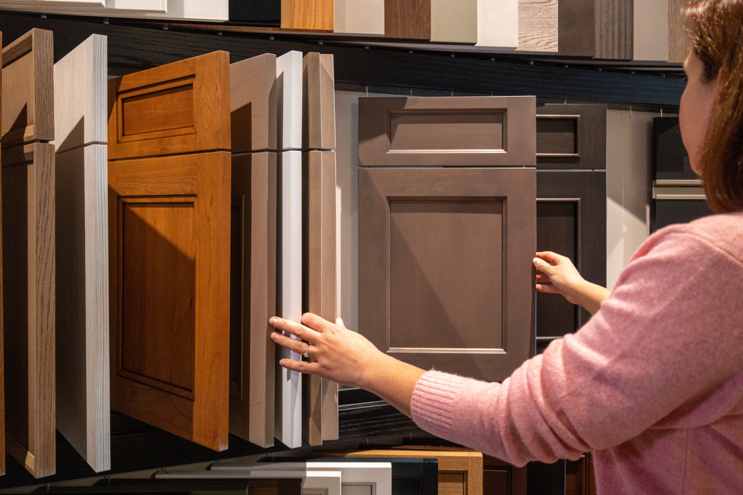 A person in a pink sweater looks at and touches various cabinet door samples displayed on a wall, exploring options for luxury interior design with the eye of an interior designer in Massachusetts.