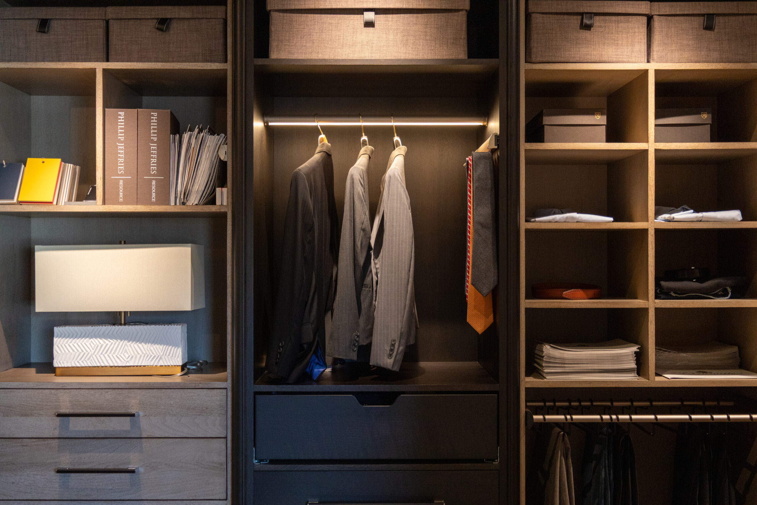 A neatly organized closet with suits, ties, boxes, folded clothes, books, a lamp, and storage shelves showcases the elegance of luxury interior design by an expert interior designer in Massachusetts.