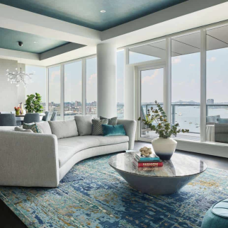 Modern living room with large windows overlooking water, a curved light gray sofa, a round marble coffee table, and blue patterned rug—a stunning example of luxury interior design by an interior designer in Massachusetts.