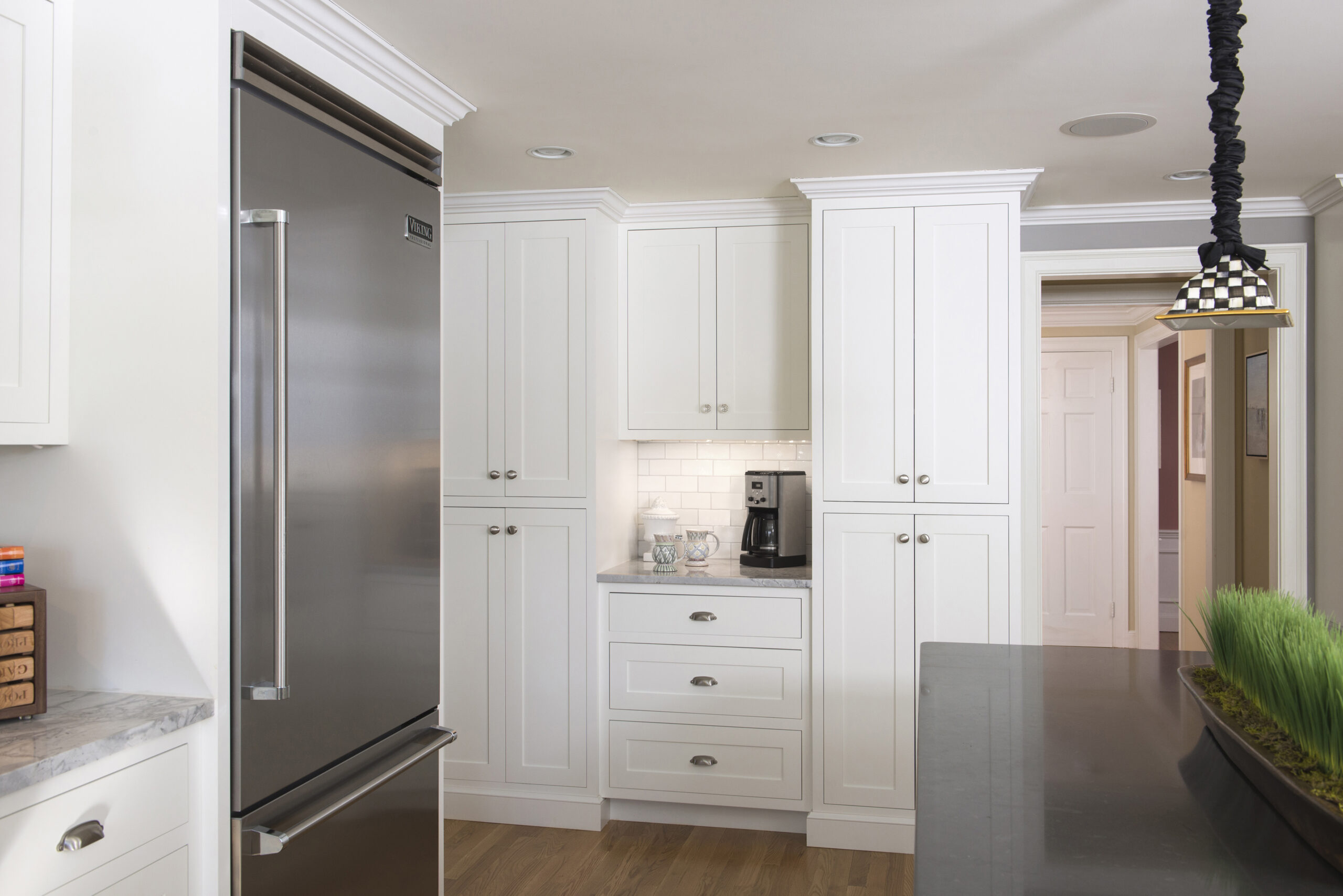 Modern kitchen with white cabinets, stainless steel refrigerator, a coffee maker on the countertop, and a checker-patterned pendant light above a dark island—showcasing luxury interior design by an expert interior designer in Massachusetts.