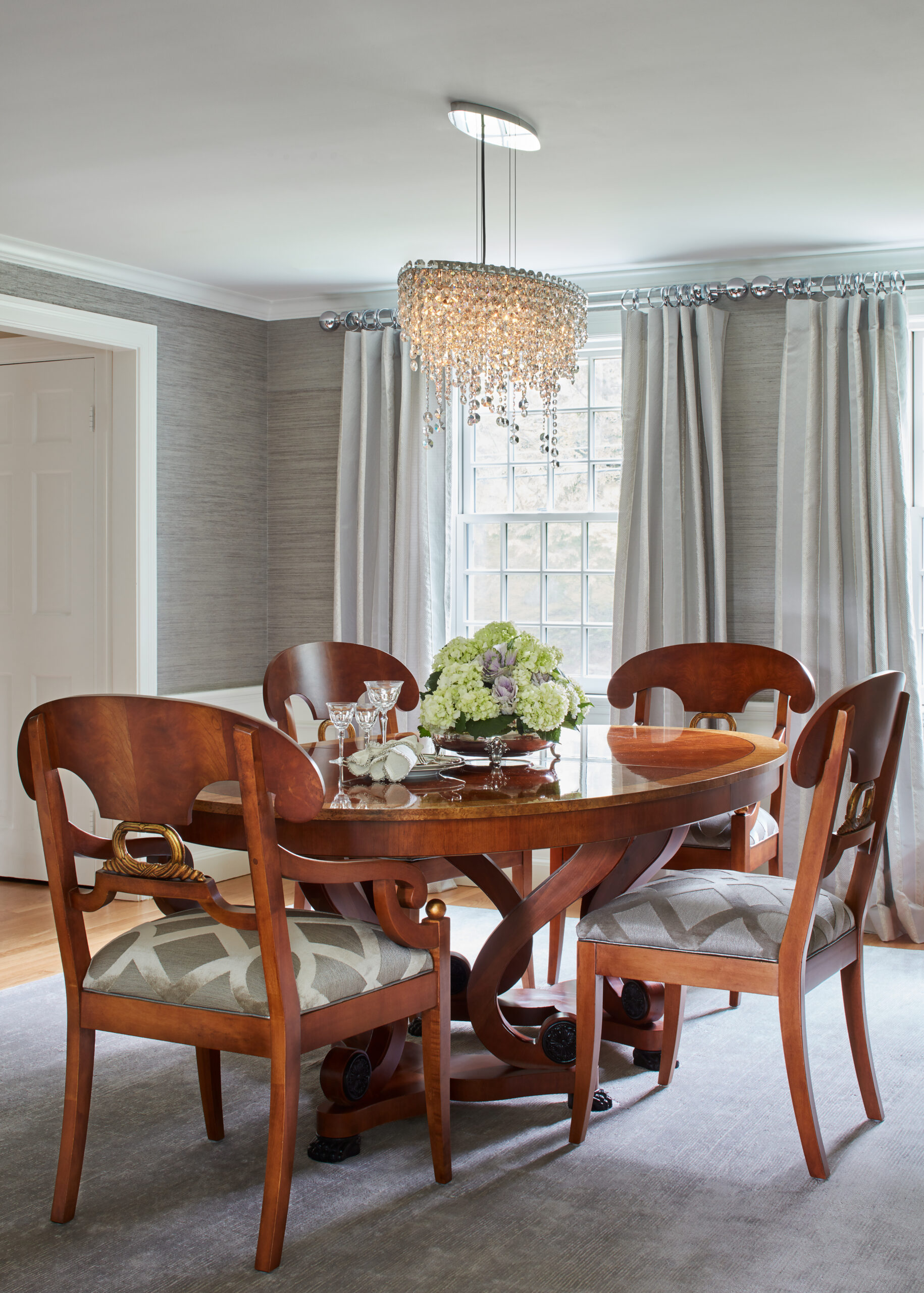 A dining room with a round wooden table, four matching chairs, a floral centerpiece, and a crystal chandelier reflects luxury interior design, set by a large window with light gray curtains.