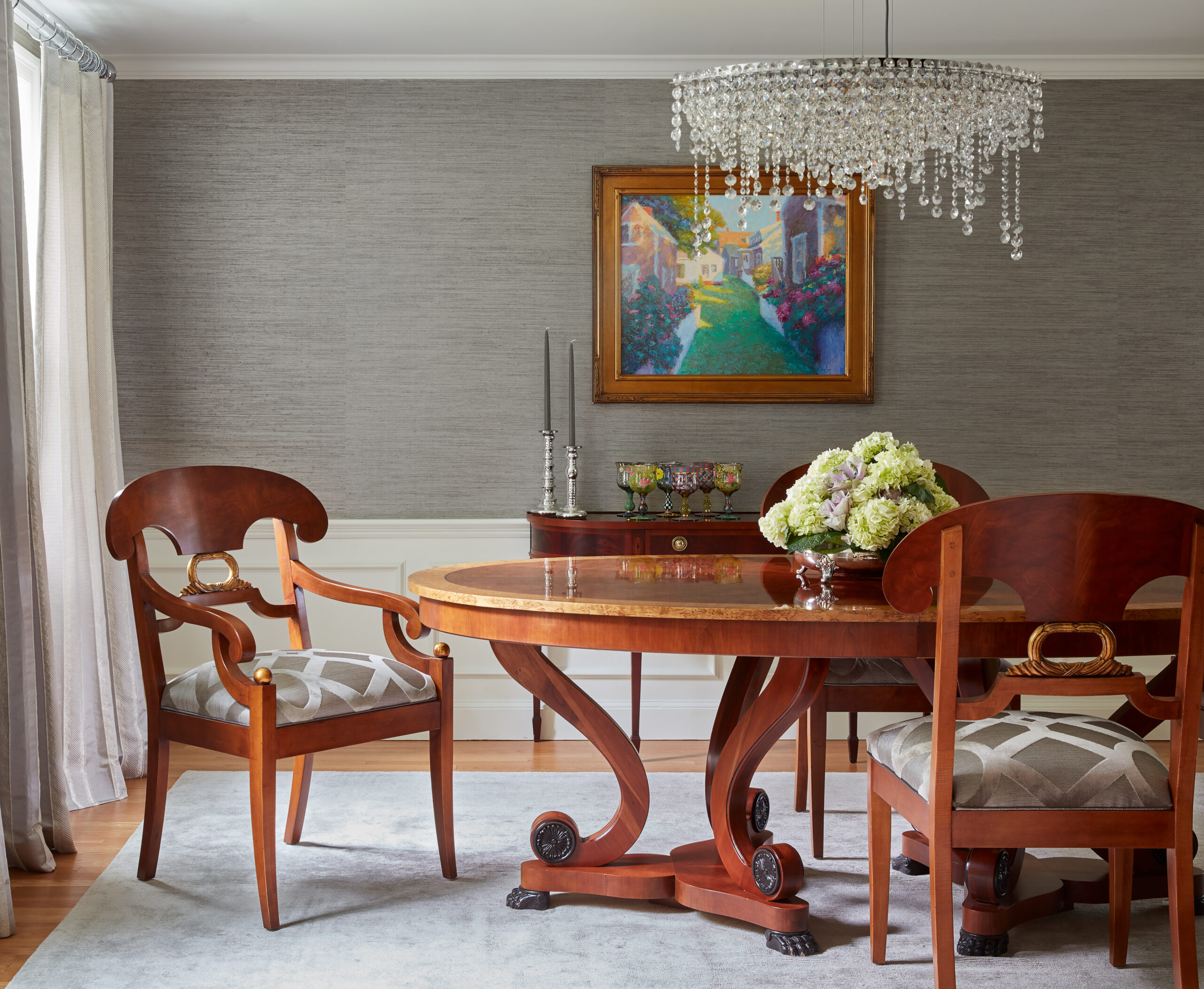 Elegant dining room with a round wooden table, four matching chairs, a crystal chandelier, and a colorful framed painting on a gray textured wall—showcasing the refined touch of Luxury Interior Design.